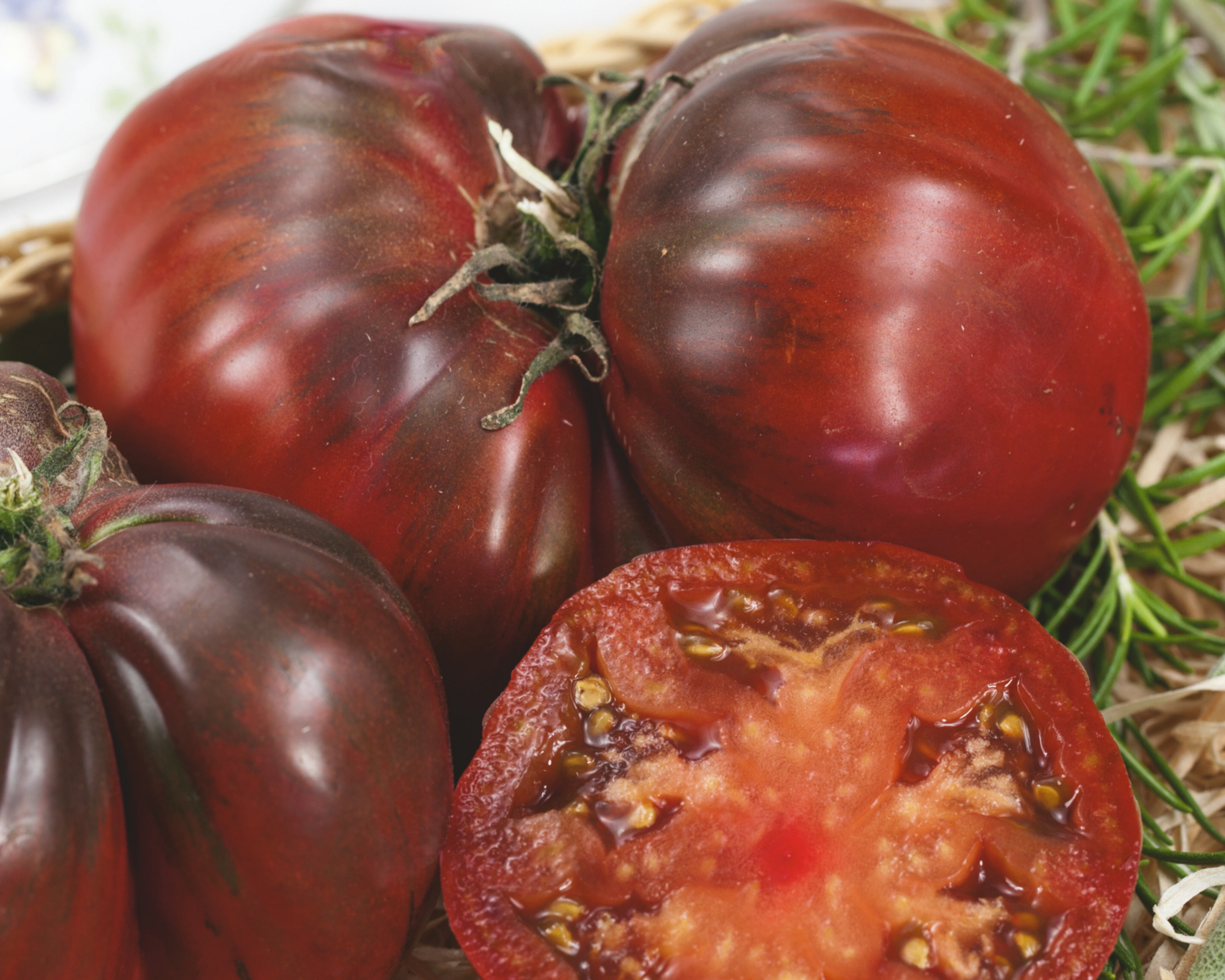 Close-up of Black Krim heirloom tomatoes from Seeds 'n Such, including one sliced open to reveal a juicy dark red-purple interior, surrounded by fresh rosemary sprigs.