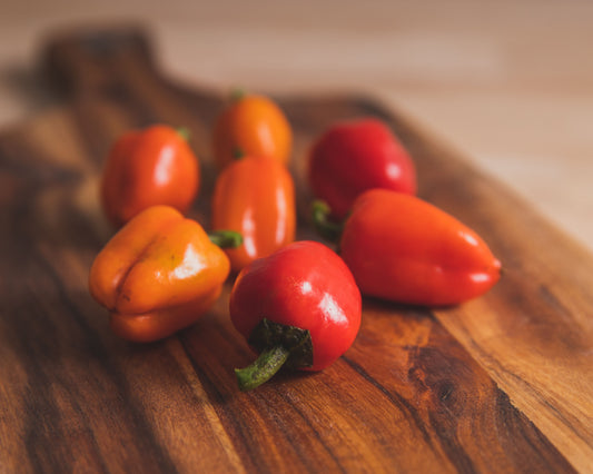 Six vibrant orange and red Mini Belle Mix peppers, grown from Seeds 'n Such seeds, are displayed on a wooden cutting board with a softly blurred background.