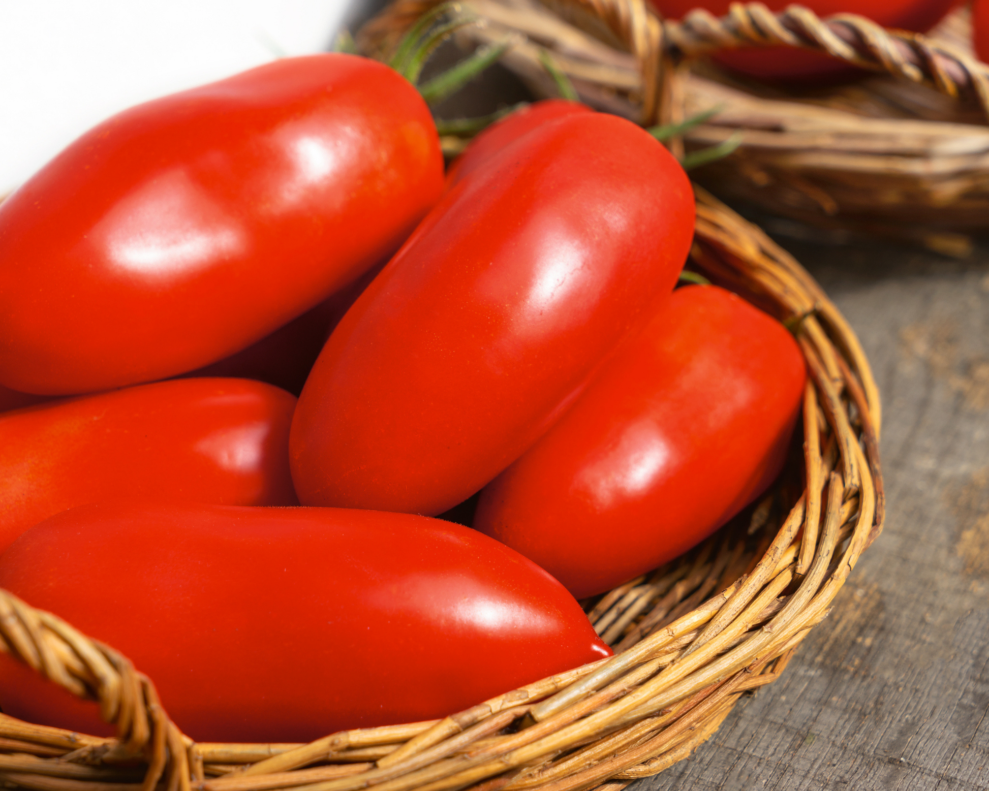 Several ripe Polish Paste tomatoes, grown from Seeds 'n Such seeds, are piled in a woven basket with another basket partly visible in the background on a rustic wooden surface.