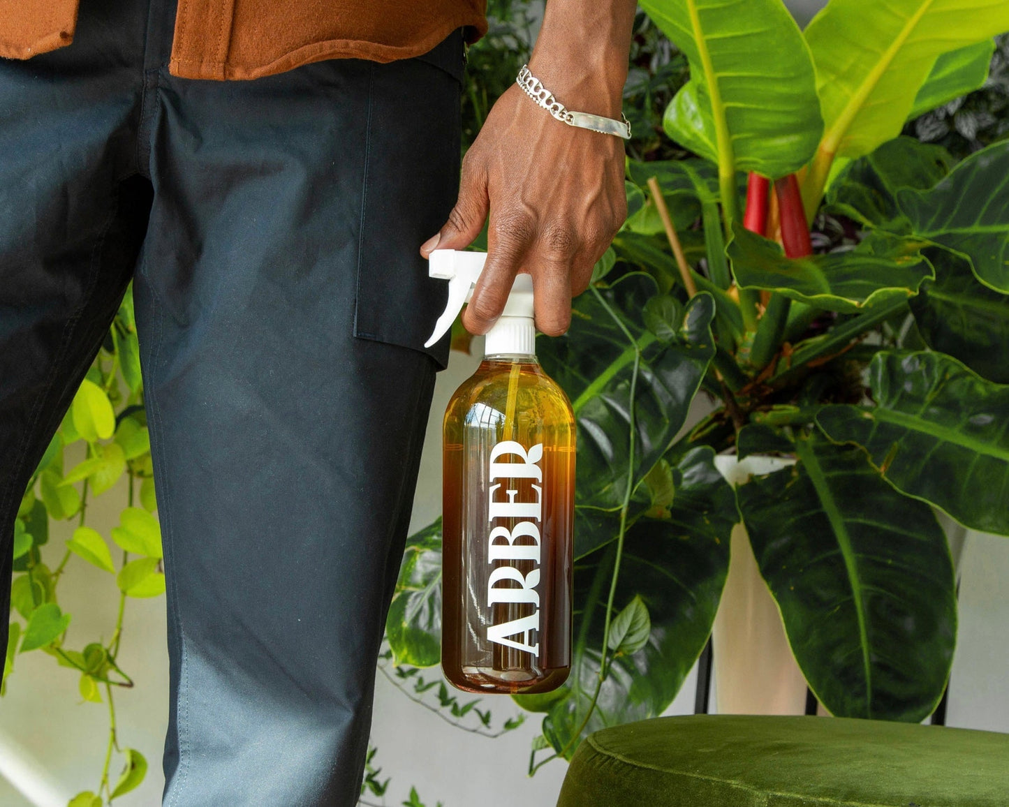A person holds a clear Arber Spray Bottle beside lush green indoor plants, dressed in dark pants and a brown top with a silver bracelet, highlighting garden hydration and plant wellness.