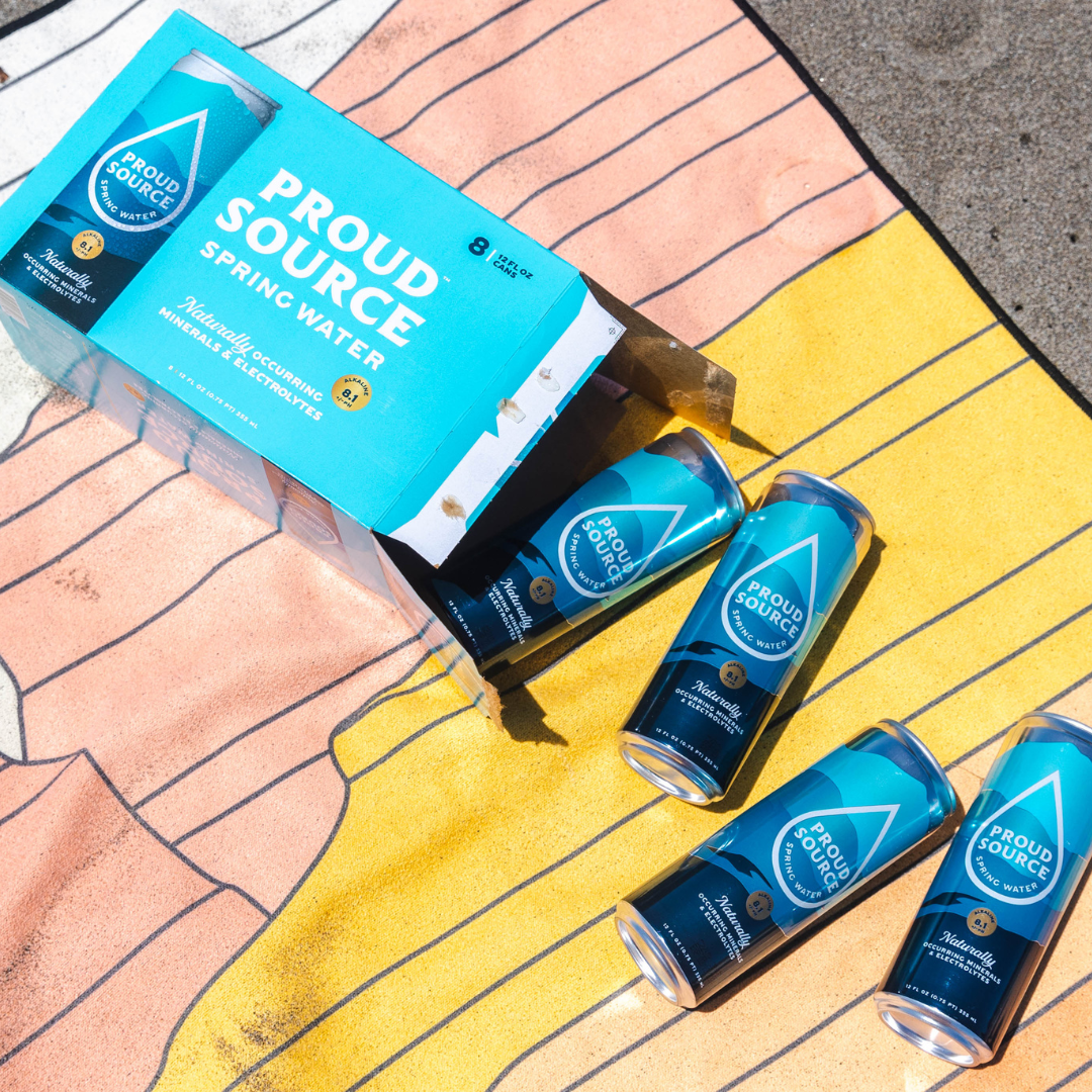 Two cans of PROUD SOURCE WATER alkaline spring water, showing the label and clear liquid inside the cans.