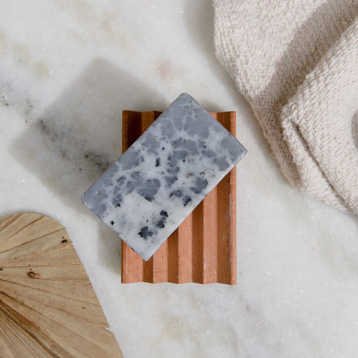 A 4oz all-natural, vegan Soap Bar from ZeroWasteStore.com sits on a wooden dish atop marble, with a beige washcloth and dried palm leaf nearby; available in 6 scents and plastic-free for face and body.