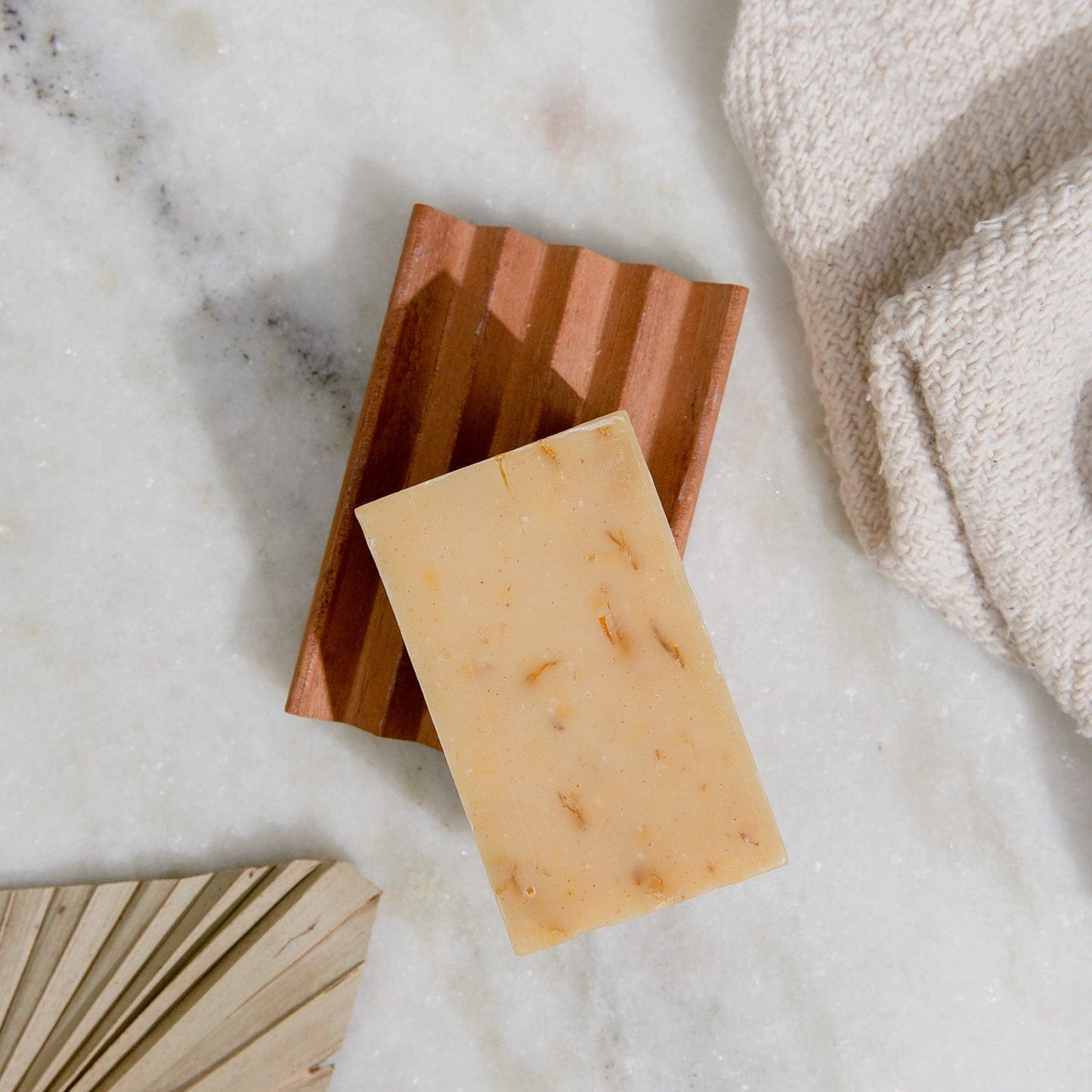 A ZeroWasteStore.com 4oz all natural, vegan soap bar for face and body—available in 6 scents—rests on a wooden dish atop marble, beside a beige towel and folded palm leaf. Plastic free.