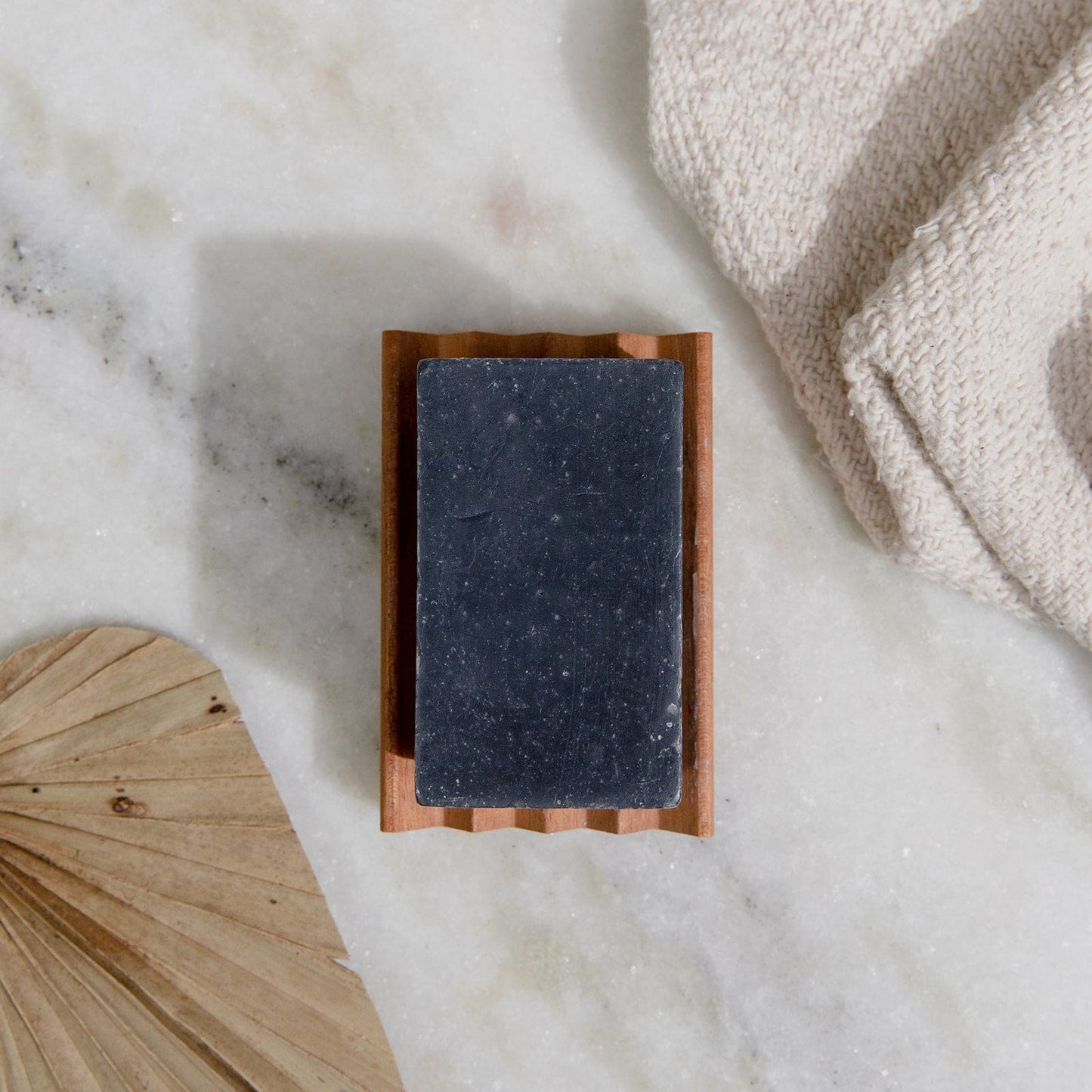 A rectangular 4oz black Soap Bar from ZeroWasteStore.com, all natural and vegan with 6 scent options, rests on a wooden dish atop marble. A folded beige towel and dried palm leaf add a spa-like touch. Plastic free packaging.