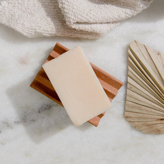 A beige 4oz all natural vegan Soap Bar from ZeroWasteStore.com rests on a wooden dish atop white marble, beside a textured towel and dried palm leaf. Available in 6 scents, it's plastic free and ideal for face and body.