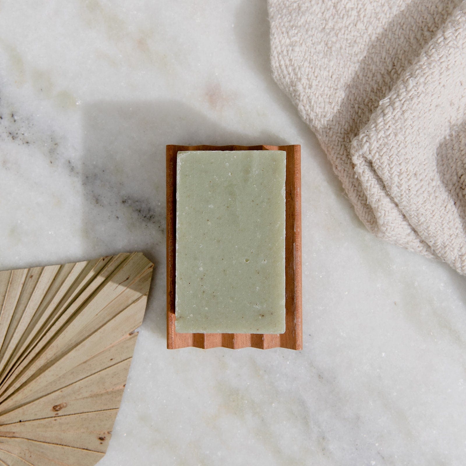 A 4oz all-natural vegan bar soap from ZeroWasteStore.com sits on a wooden dish atop marble, with a beige towel and palm leaf nearby. Plastic-free and available in 6 scents for face and body use.