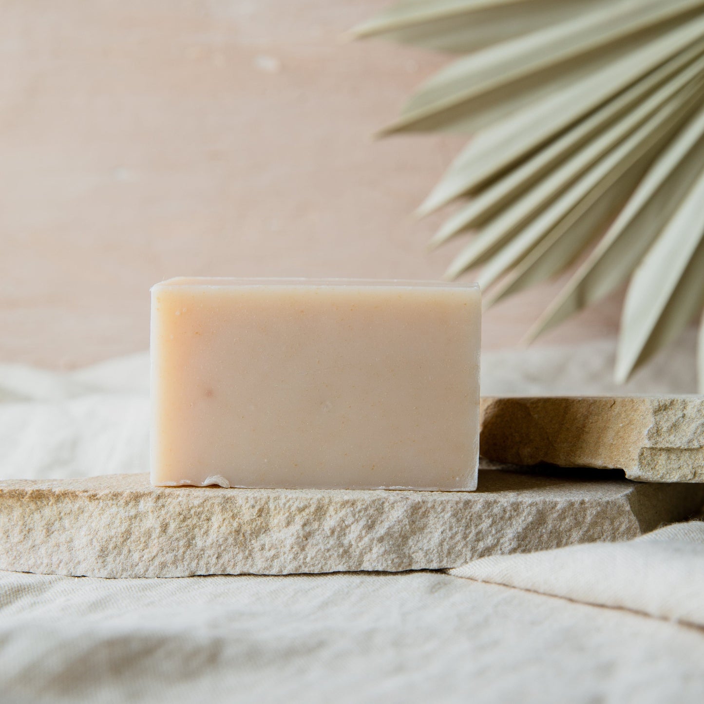 A beige 4oz all-natural vegan soap bar from ZeroWasteStore.com sits on stacked stone slabs with a blurred dried palm leaf behind and neutral fabric underneath. Available in 6 scents; plastic-free and perfect for face or body.