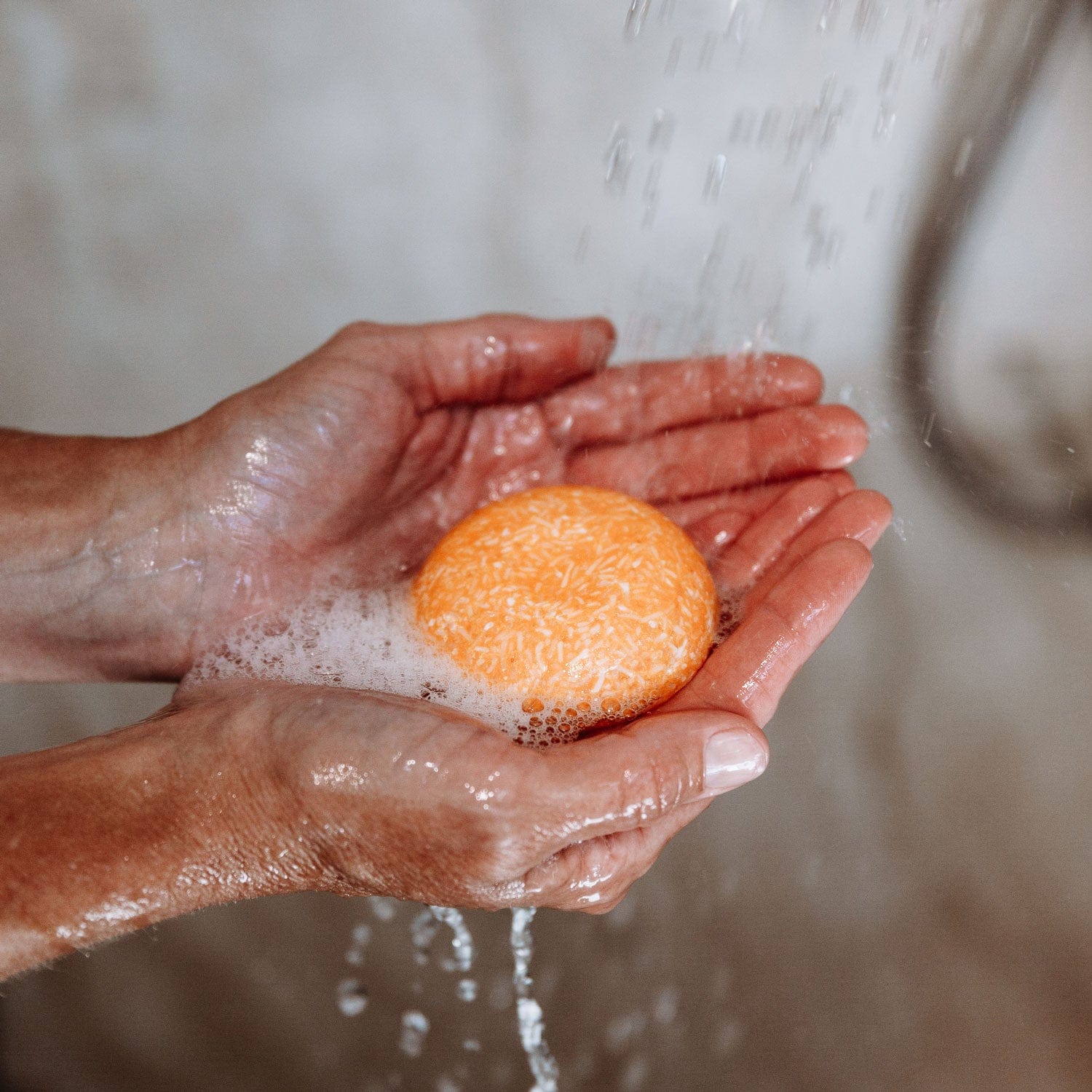 A person’s wet hands hold a round orange ZeroWasteStore.com Shampoo Bar under running water, creating lather and bubbles. The background is blurred.