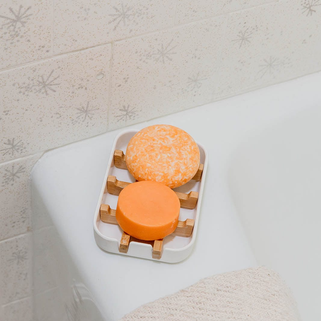 Two orange Shampoo & Conditioner Bar Duo bars from ZeroWasteStore.com sit on a white and wooden soap dish by a bathtub, against textured tile—ideal for a zero waste bathroom routine.