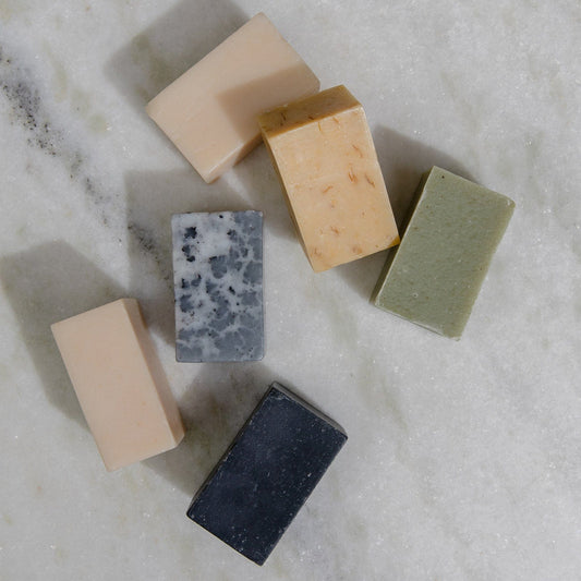 Six all-natural, vegan 4oz soap bars from ZeroWasteStore.com—in beige, green, black, speckled gray, and tan—are arranged on white marble. Each bar offers a unique scent and is crafted for face and body with plastic-free packaging.