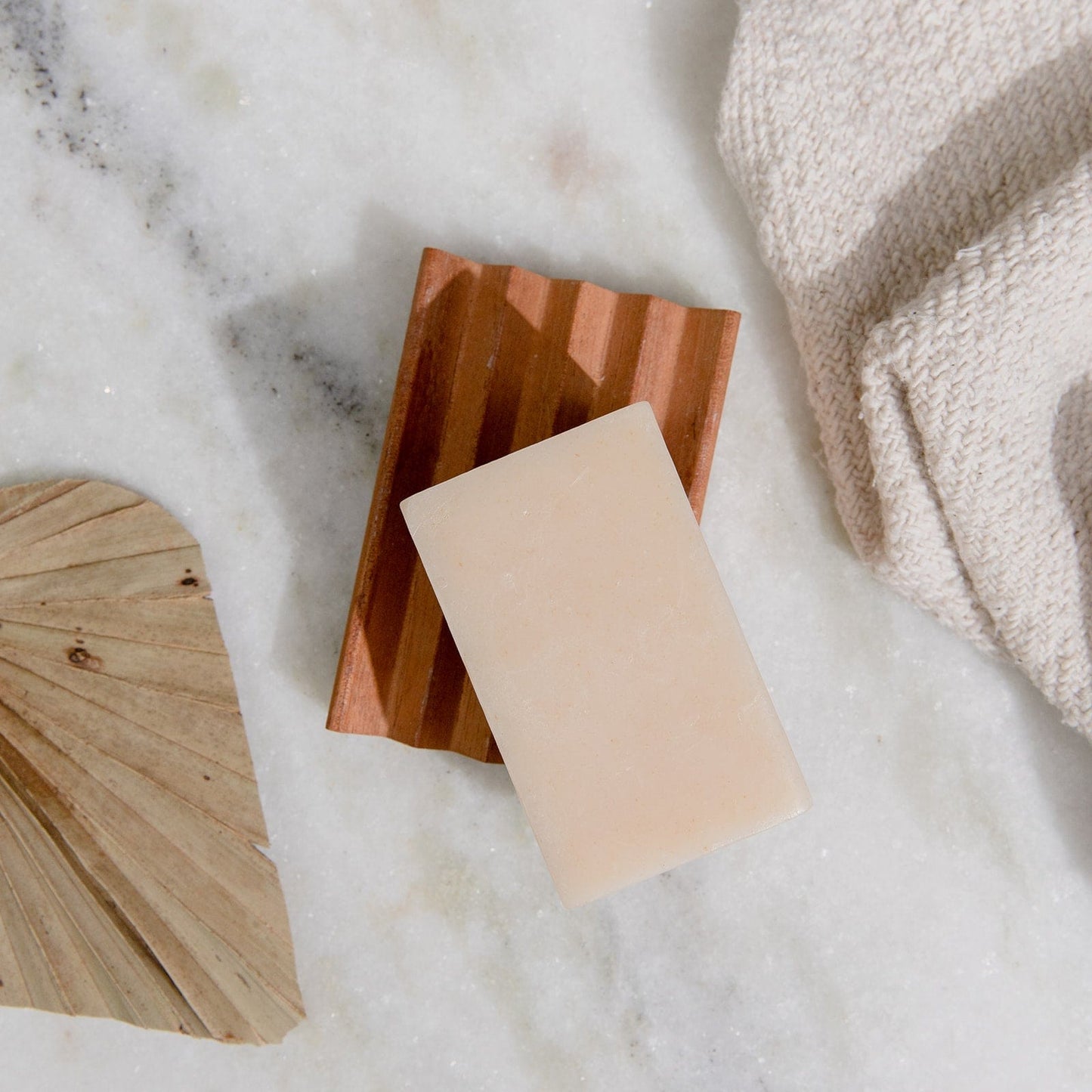 A 4oz all-natural, vegan Soap Bar from ZeroWasteStore.com sits on a wooden dish atop marble, beside a beige towel and dried palm leaf. Choose from 6 plastic-free face and body scents.