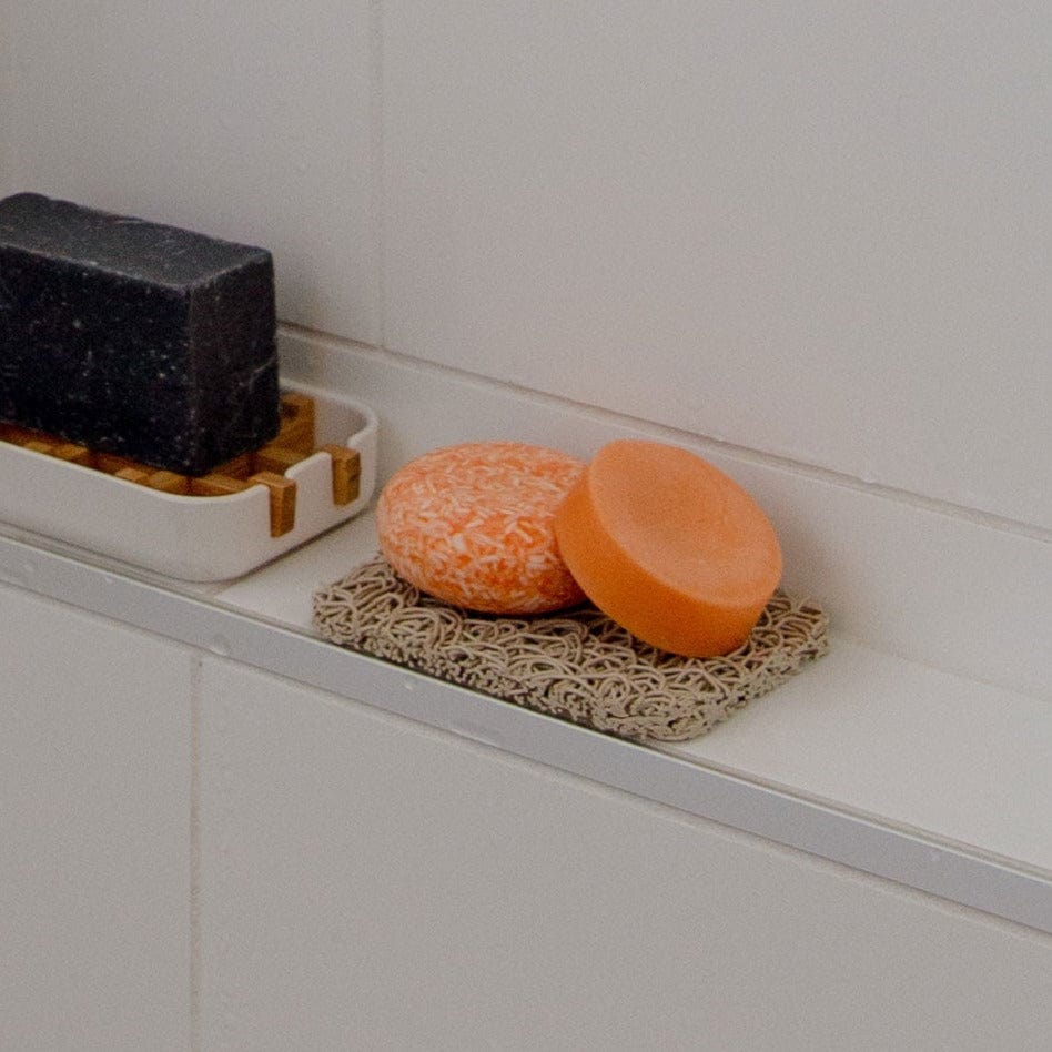 Two orange round soap bars and a black rectangular bar sit on separate ZeroWasteStore.com Soap Rest holders, made from corn-based plastic, displayed on a bathroom ledge against a white tiled wall.