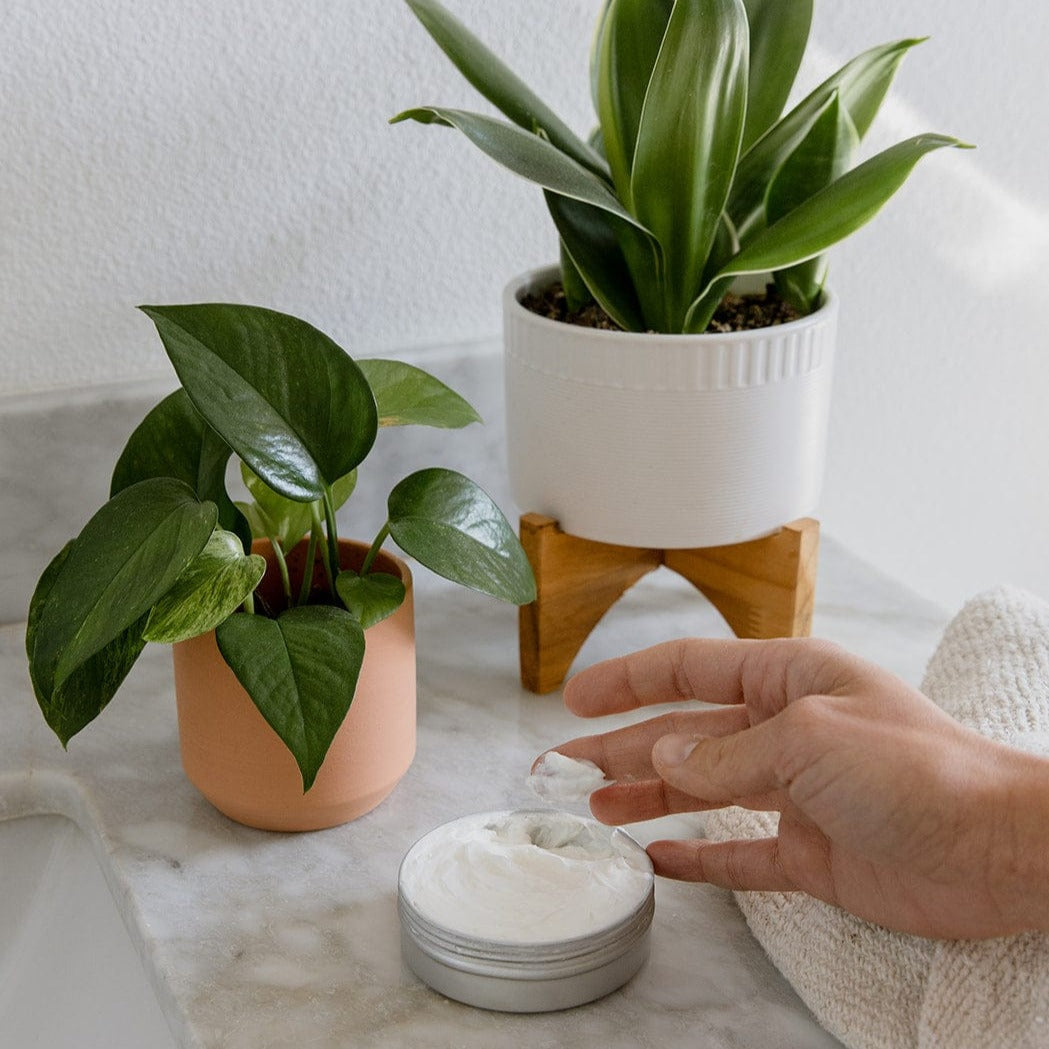 A hand scoops ZeroWasteStore.com’s Zero Waste Body Butter (Shampoo Bar Scents, 4oz) from a round tin on a marble countertop, beside two houseplants—one in a peach pot and one in a white pot with a wooden stand.