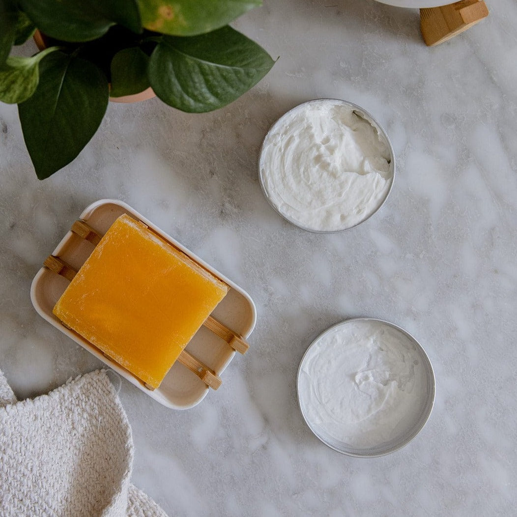 A yellow soap bar and two open tins of ZeroWasteStore.com Zero Waste Body Butter (Shampoo Bar Scents, 4oz, Deeply Nourishing, Moisturizing) rest on marble beside a textured cloth and leafy plant.