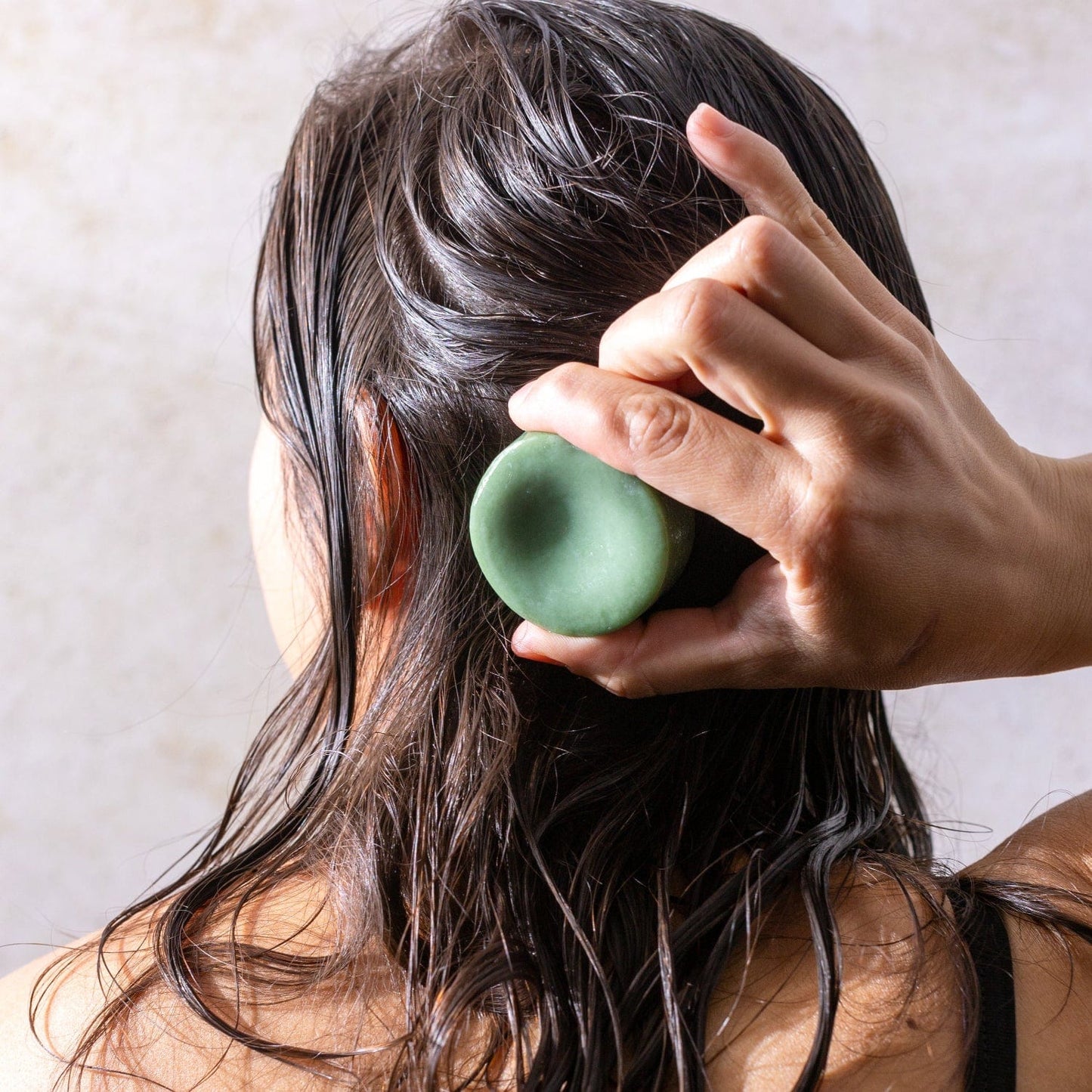 A person with wet, dark hair is seen from behind rubbing the Bloom Castor Oil Conditioner Bar by ZeroWasteStore.com onto their head for stronger, softer, healthier hair and plastic-free care.