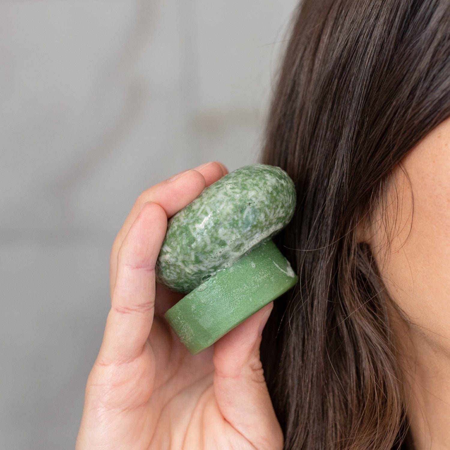 A person with long brown hair holds two Bloom Castor Oil Conditioner Bars by ZeroWasteStore.com near their head, highlighting plastic-free hair care for stronger, softer, healthier hair against a neutral background.