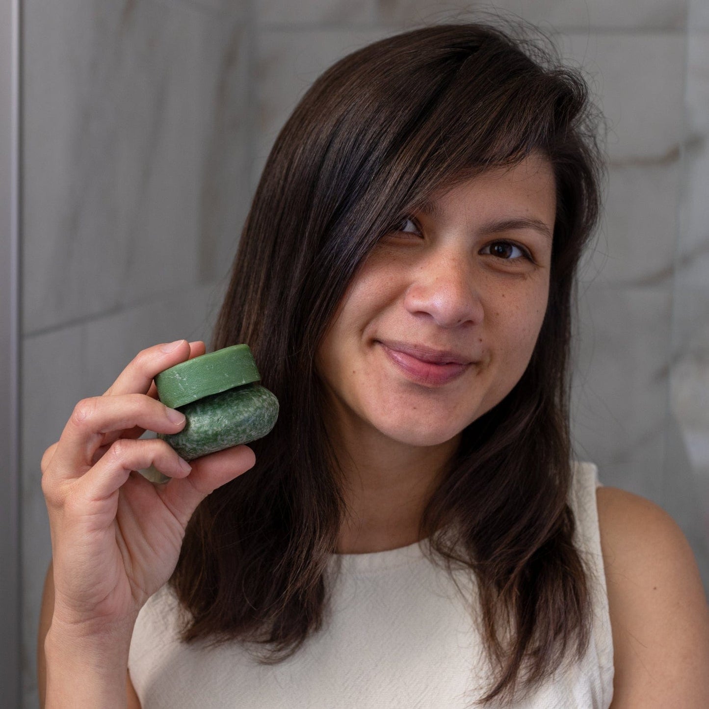 A woman with long dark hair and a white sleeveless top smiles while holding up the Bloom Castor Oil Shampoo Bar for Hair Growth & Scalp Revival from ZeroWasteStore.com in a tiled bathroom.