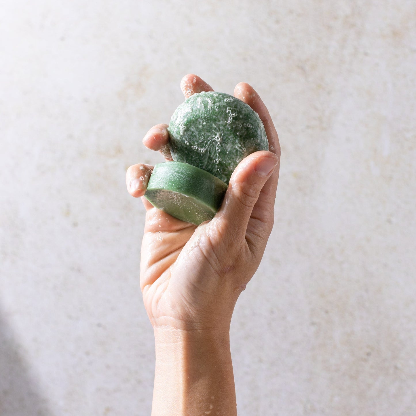 A hand holds the Bloom Shampoo & Conditioner Bar Duo by ZeroWasteStore.com, a green, round bar with bubbles—infused with castor oil to boost scalp health and support hair growth.