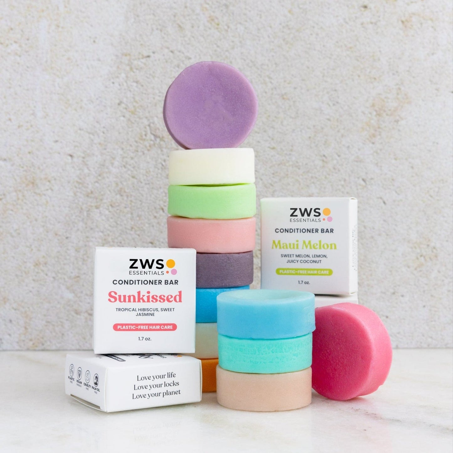 Colorful round shampoo bars and ZeroWasteStore.com sulfate-free Conditioner Bars are stacked with two ZWS Essentials boxes labeled "Sunkissed" and "Maui Melon," all arranged on a light surface against a neutral background.