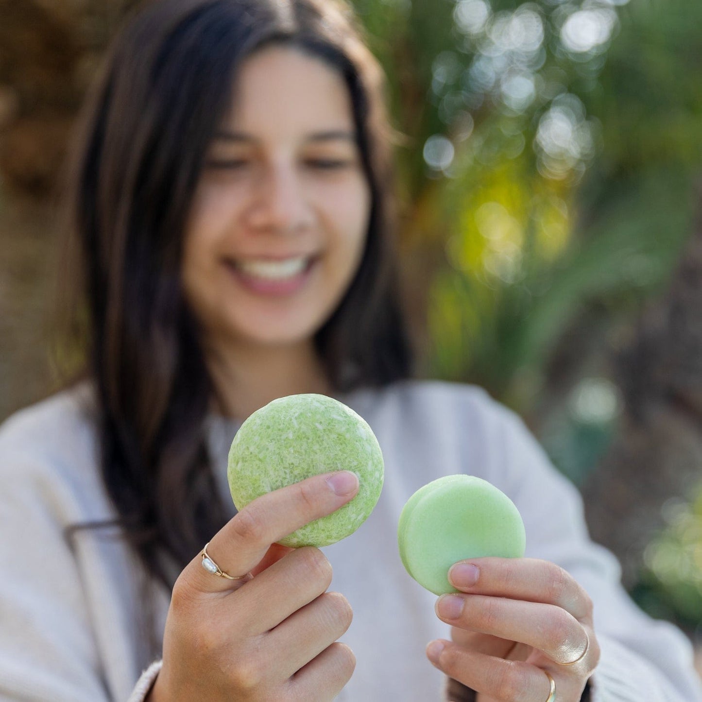 A woman with long dark hair smiles outdoors, holding a green macaron and the ZeroWasteStore.com Conditioner Bar, comparing their sizes to showcase this sulfate free bar as a sustainable choice for zero waste living.