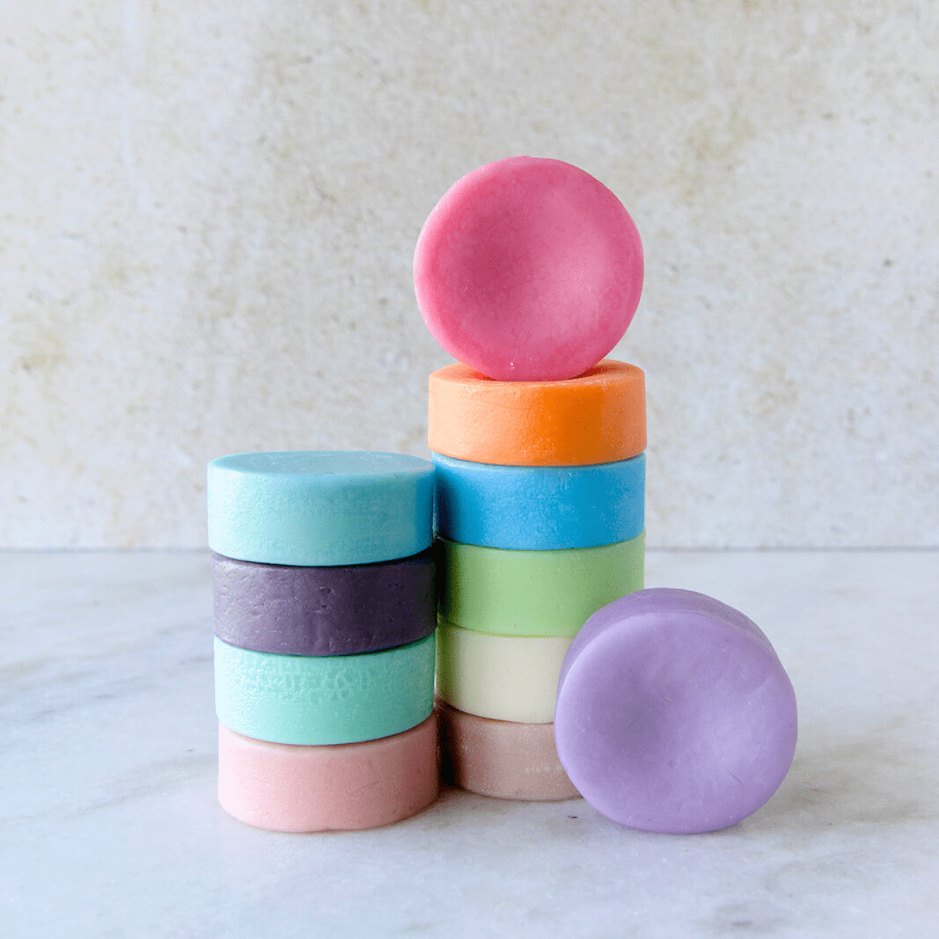 A stack of colorful pastel sulfate-free soaps is arranged on a light surface, with one pink soap on top and the ZeroWasteStore.com Conditioner Bar placed in front.
