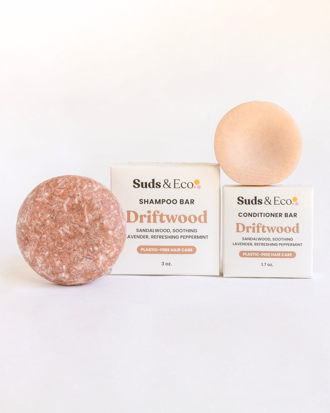 Two round bars, one reddish-brown and one beige, sit before white ZeroWasteStore.com boxes. The Shampoo & Conditioner Bar Duo features sandalwood, lavender, and peppermint scents in a zero waste formula.