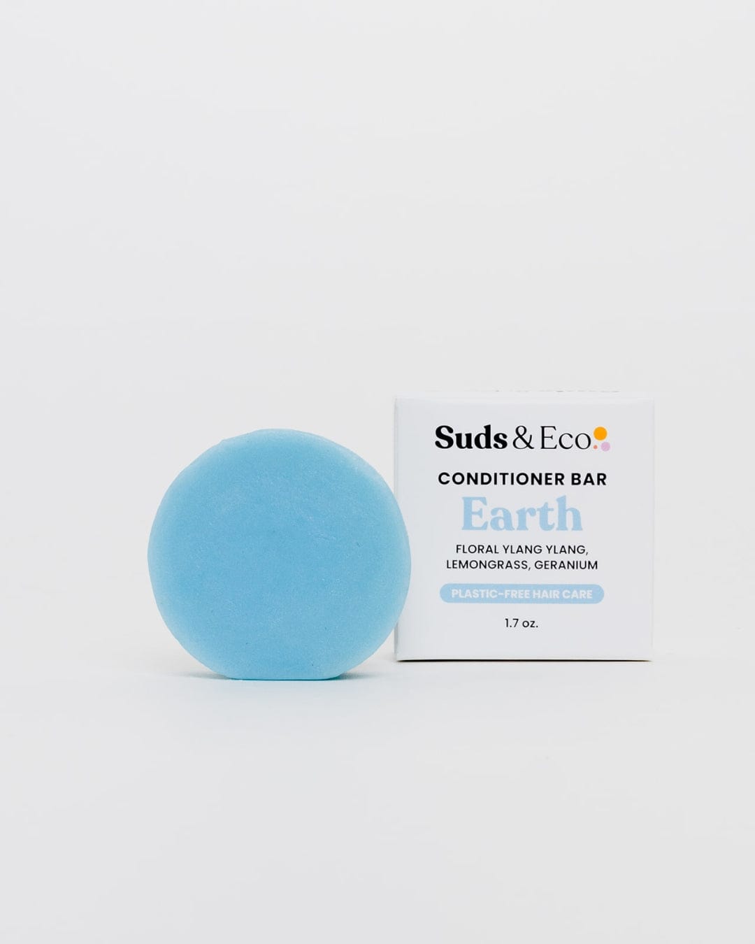 A round, light blue Conditioner Bar from ZeroWasteStore.com rests by its white, branded packaging. This plastic-free bar offers floral ylang ylang, lemongrass, and geranium notes for eco-friendly hair care.