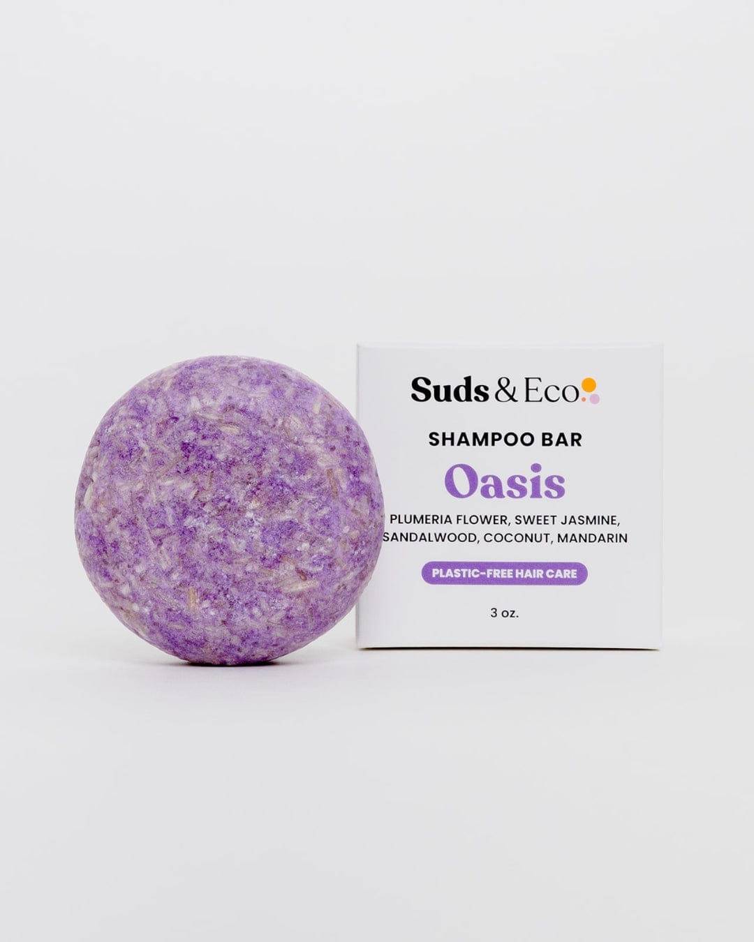 A round, purple Shampoo Bar from ZeroWasteStore.com sits next to a white box. Oasis scent has plumeria, jasmine, sandalwood, coconut, and mandarin notes; 3 oz. zero waste hair care for a plastic-free routine.