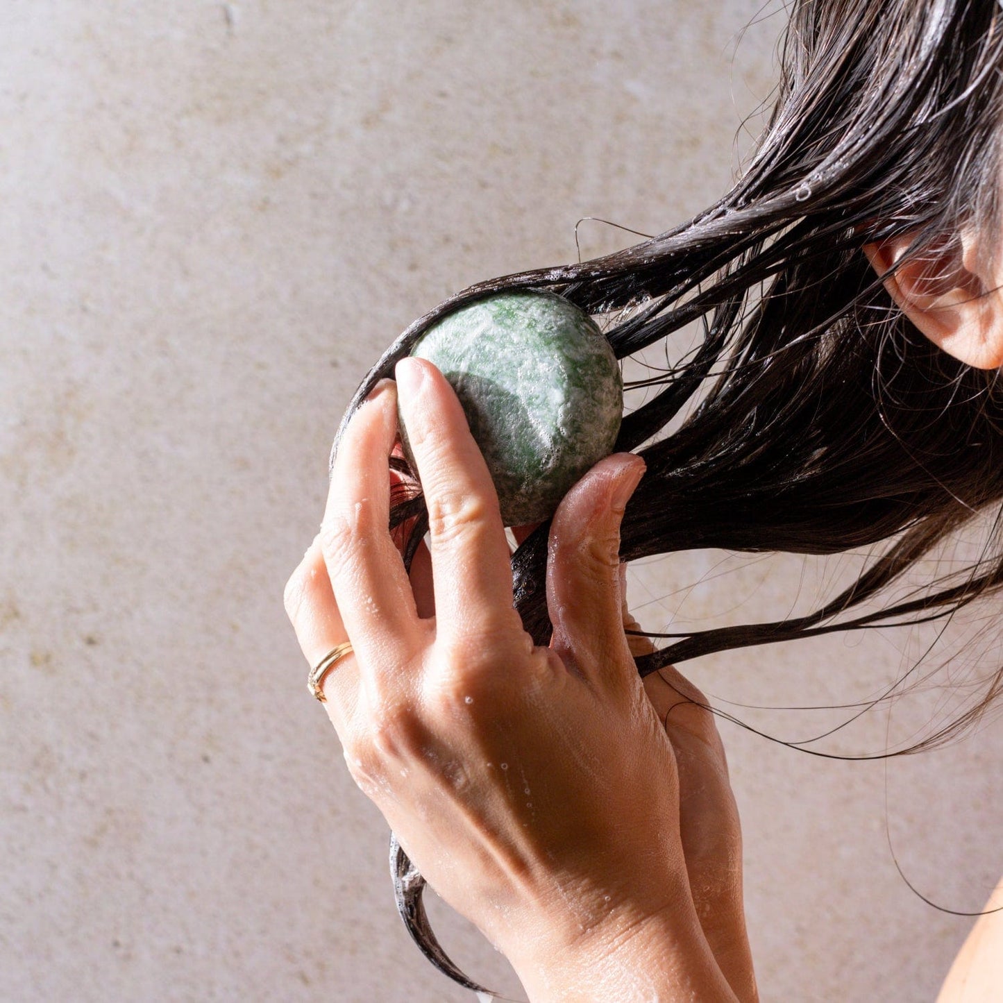 A person with wet hair lathers their hair using both hands and the green Shampoo Bar by ZeroWasteStore.com, set against a neutral background and showcasing an eco-friendly hair care routine.