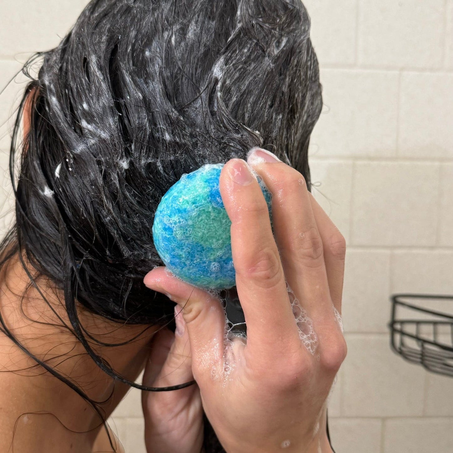 A person with wet, dark hair uses the ZeroWasteStore.com Shampoo Bar in the shower, creating a lather on their scalp. White tiled walls and a metal shower caddy appear in the background.