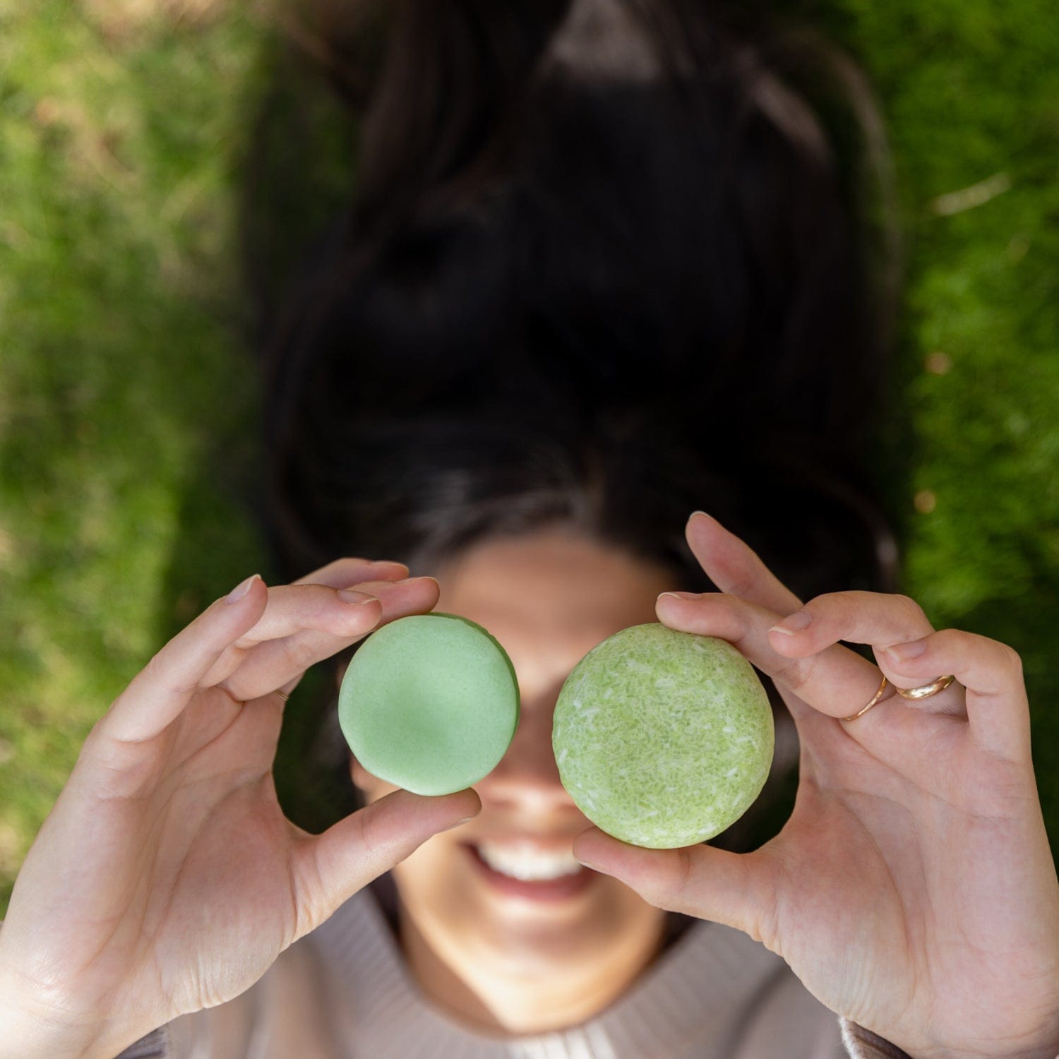 Lying on grass, a person with long dark hair holds a green macaron and a round green Shampoo Bar from ZeroWasteStore.com in front of their smiling face.