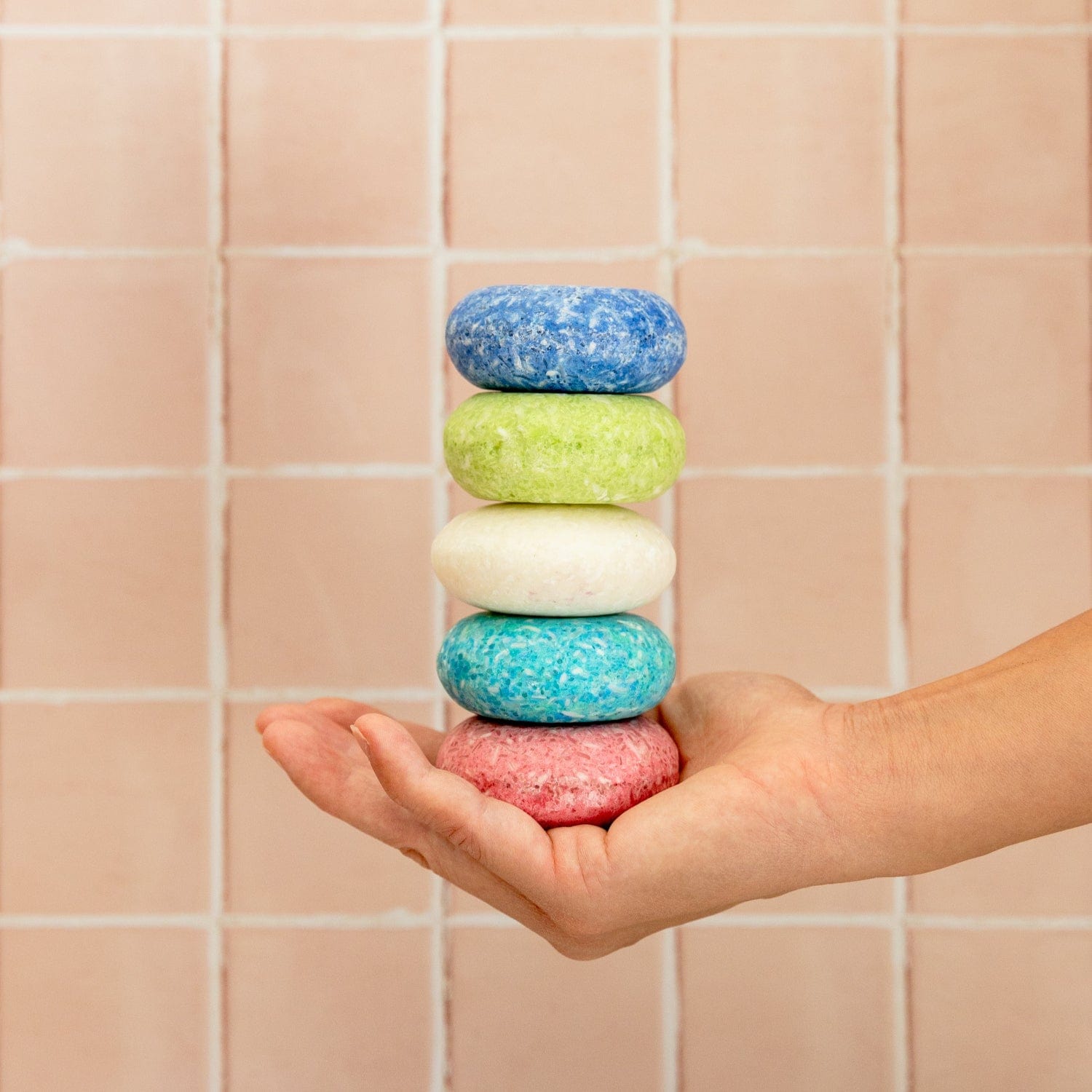 A hand holds five vibrant Shampoo Bars—blue, green, white, turquoise, and pink—from ZeroWasteStore.com against a pink tiled wall, highlighting a colorful and eco-friendly zero-waste hair care option.