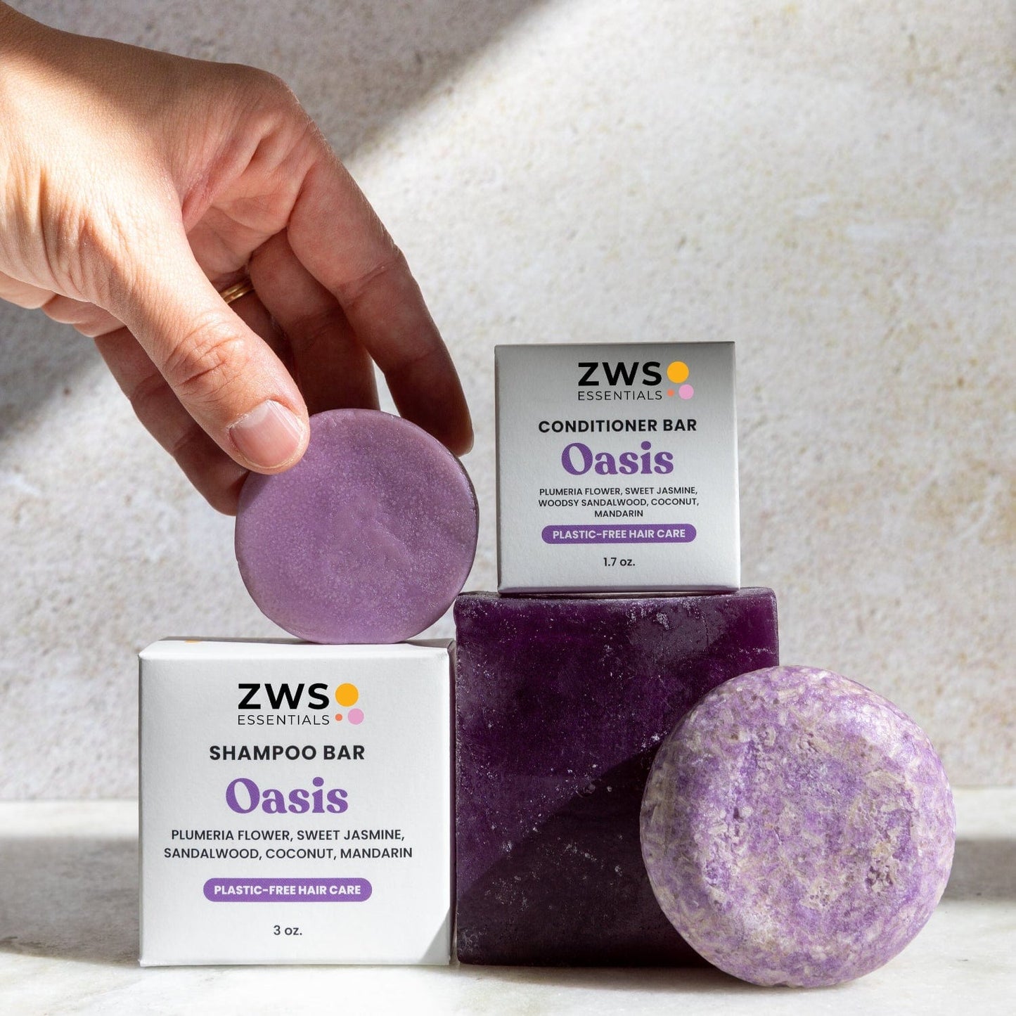A hand holds a round purple Shampoo & Conditioner Bar Duo by ZeroWasteStore.com, with both bars and boxes nearby, offering floral and citrus scents for plastic-free hair care.
