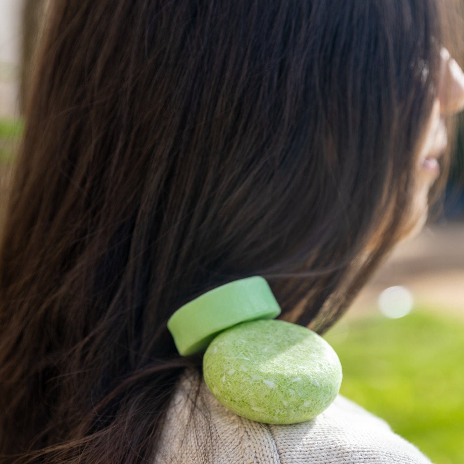 A woman with long dark hair has two solid green ZeroWasteStore.com Shampoo & Conditioner Bar Duo bars resting on her shoulder, set against a soft outdoor background.