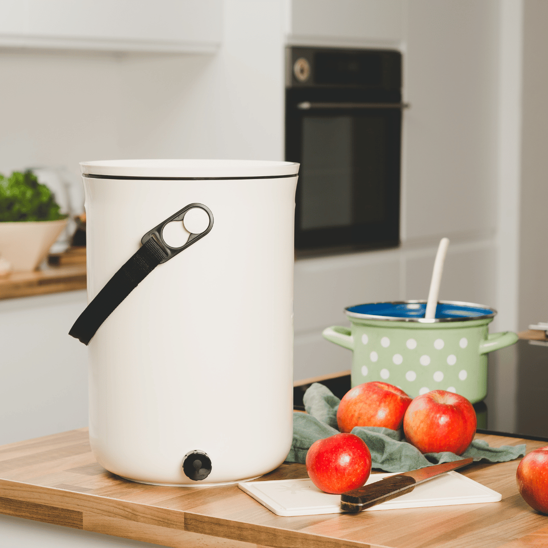 The TeraGanix Bokashi Kitchen Compost Bin, 2.5 gal, with a black handle, sits on the counter beside a cutting board, three apples, a knife, and a green polka-dot pot—perfect for modern kitchen composting.