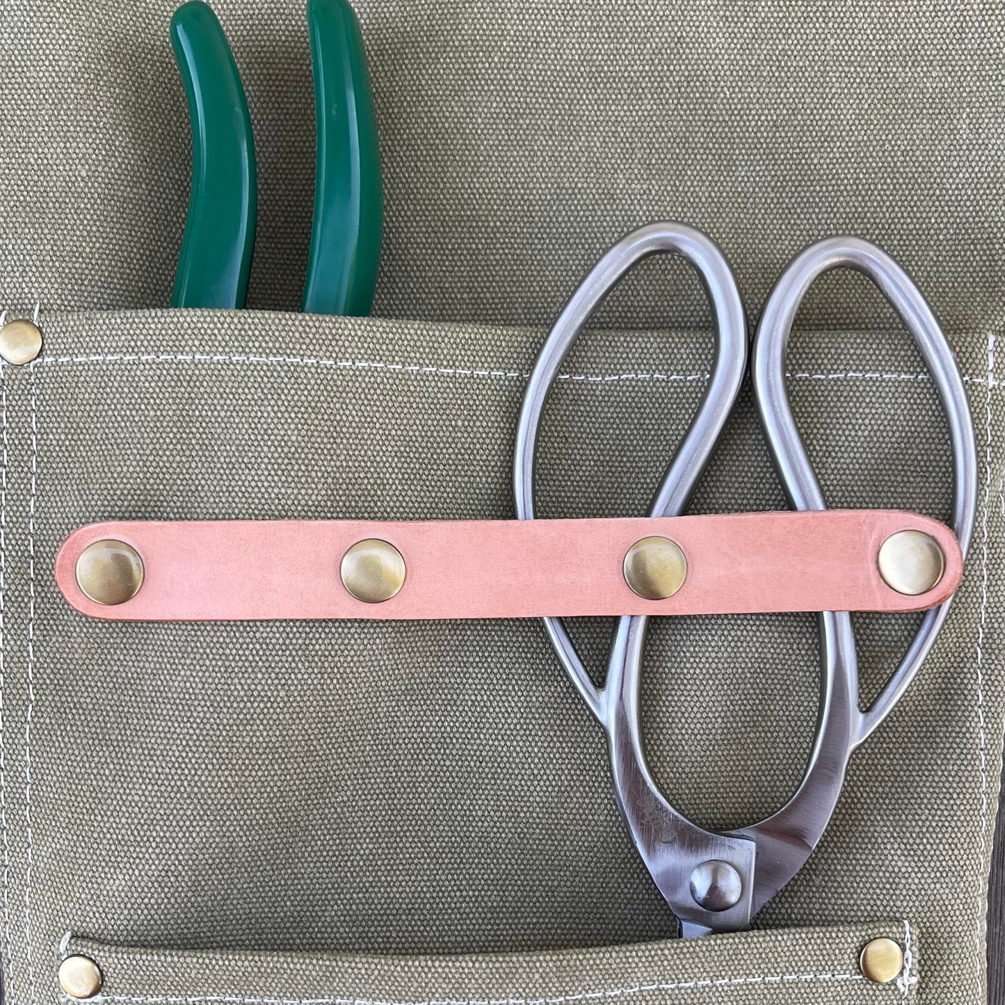 The Celtic Farm Gardening Apron—waxed canvas with white stitching, brass rivets, and roomy pockets—holds green-handled pliers and silver scissors with a leather strap, ideal for florists seeking style and durability.
