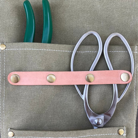 The Celtic Farm Gardening Apron—waxed canvas with white stitching, brass rivets, and roomy pockets—holds green-handled pliers and silver scissors with a leather strap, ideal for florists seeking style and durability.