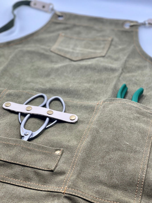The Celtic Farm Gardening Apron - Waxed Canvas Apron with Pockets is shown close-up on a light surface, holding metal garden shears with a white strap and green-handled pliers in its pockets.