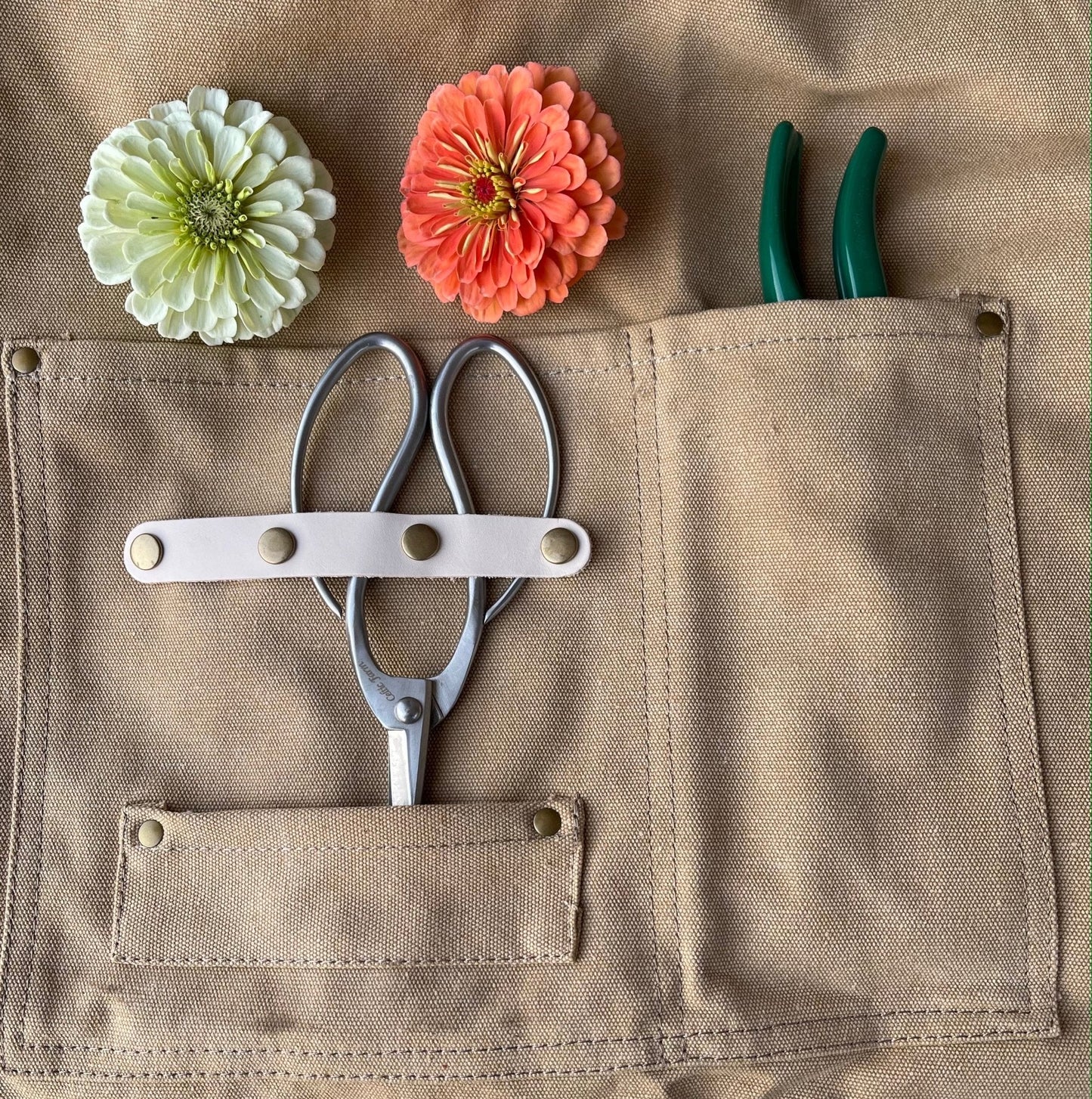 The Celtic Farm Gardening Apron—Waxed Canvas Apron with Pockets—holds zinnias, silver shears with a strap, and green-handled pruners, making it ideal for florists or gardening enthusiasts.
