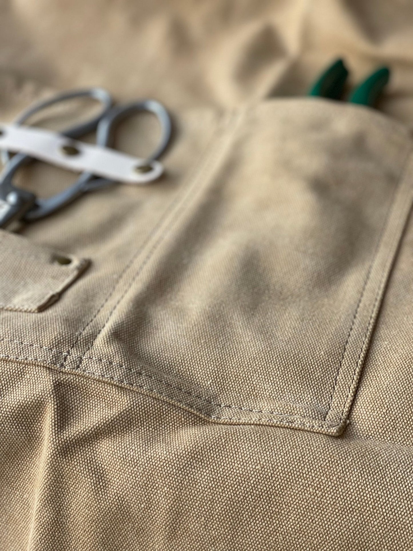 Close-up of The Celtic Farm Gardening Apron - Waxed Canvas Apron with Pockets, featuring green-handled pliers and metal scissors partially visible in a pocket on the left side.