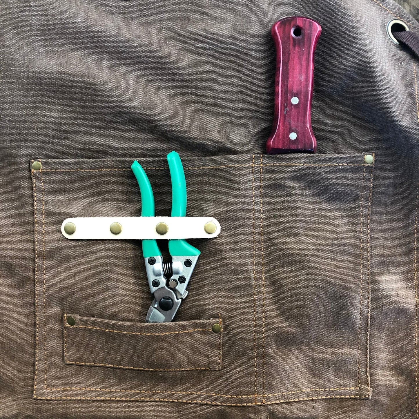 The Celtic Farm Gardening Apron – Waxed Canvas Apron with Pockets, shown in brown, holds a green-handled wire cutter in the pocket and a red-handled screwdriver tucked into a narrow slot above.
