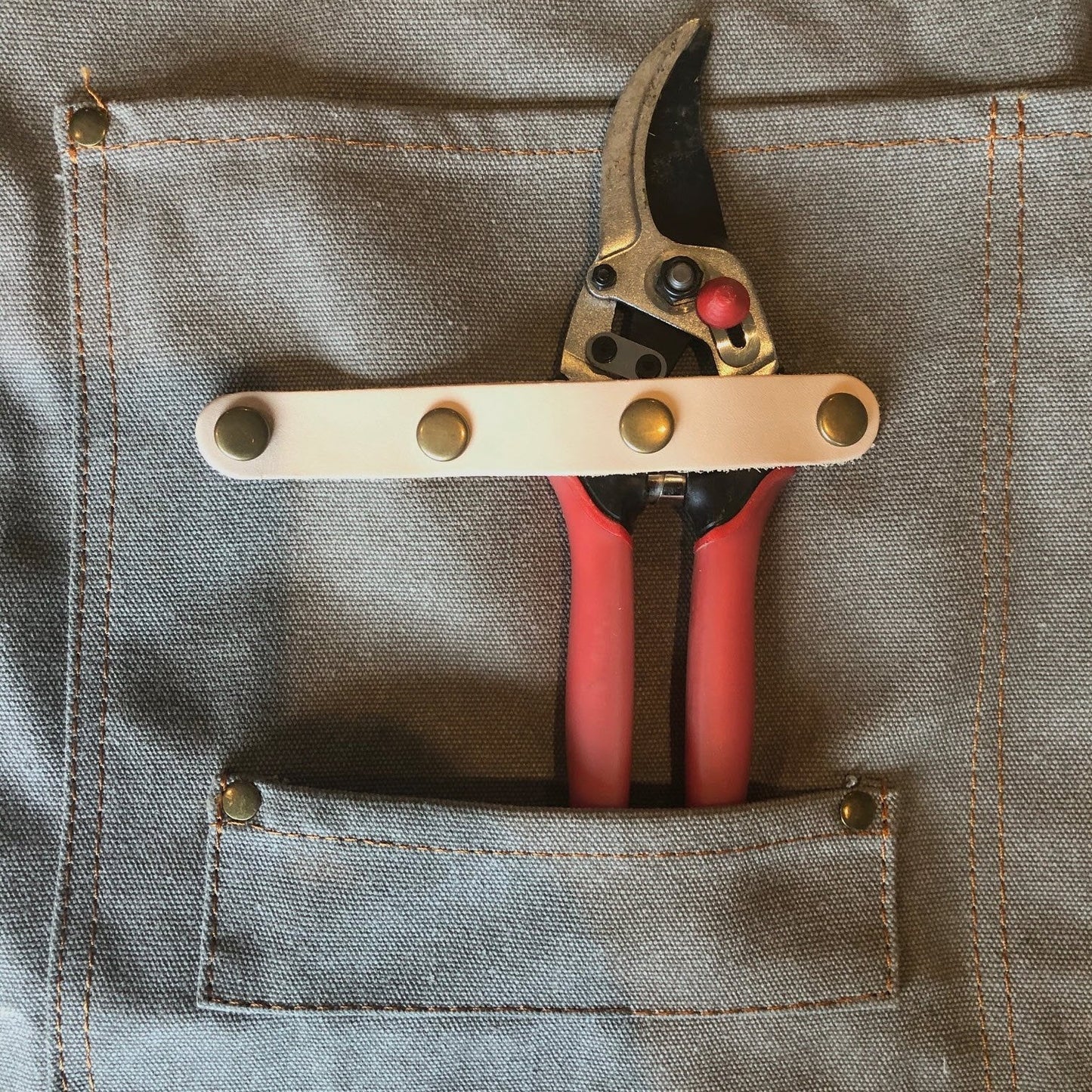 A pair of red-handled pruning shears with a leather strap is tucked into a pocket of The Celtic Farm Gardening Apron, a grey waxed canvas apron with pockets.
