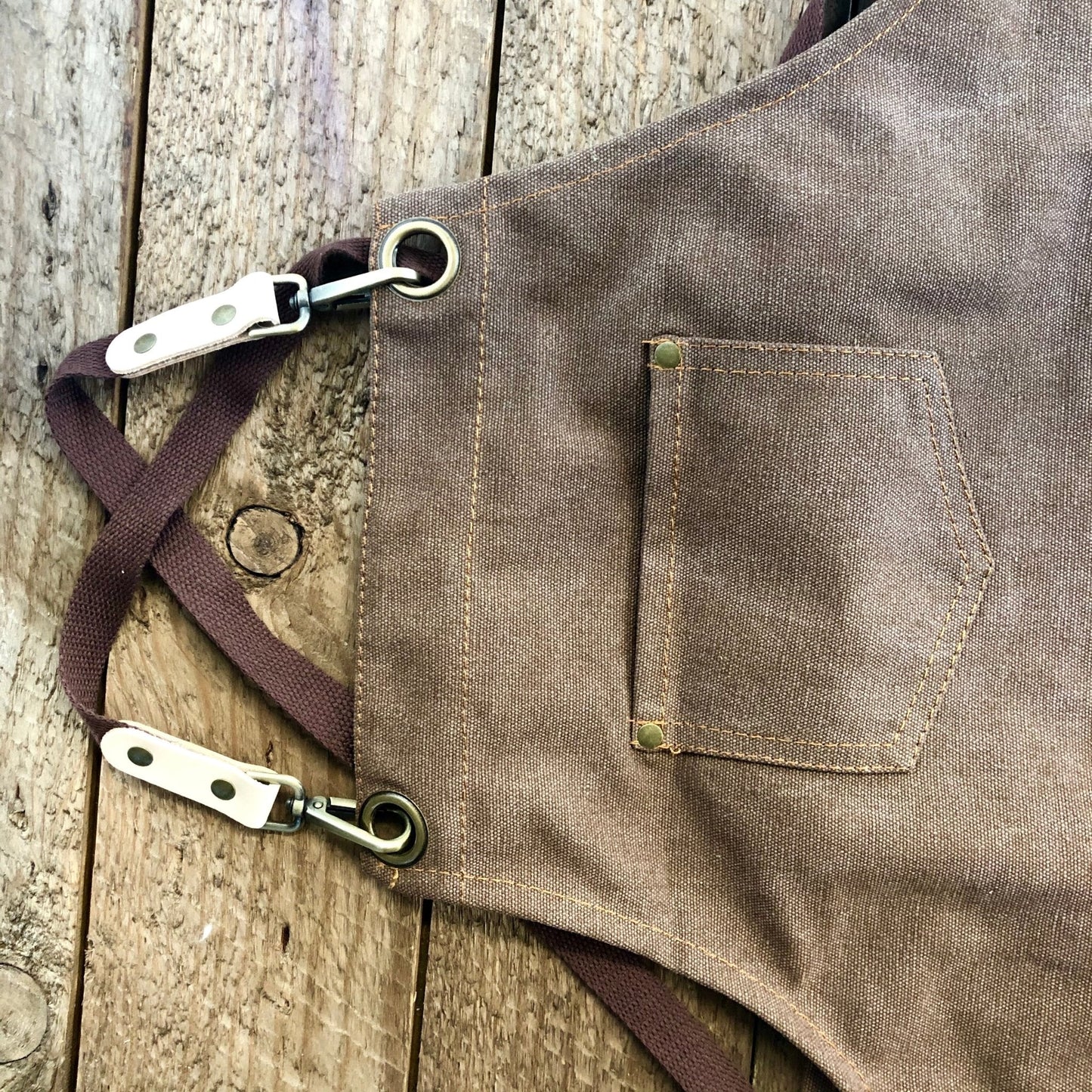 A close-up of The Celtic Farm Gardening Apron—Waxed Canvas Apron with Pockets—featuring metal clasps, a small chest pocket, and crossed straps, laid on a rustic wooden surface.