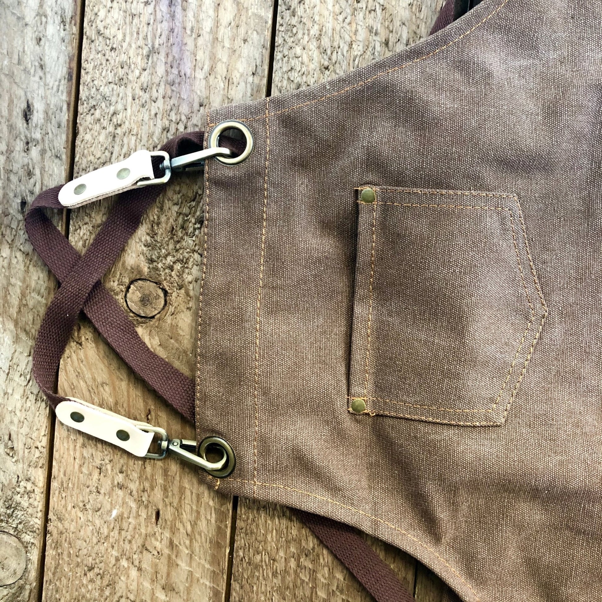 A close-up of The Celtic Farm Gardening Apron—Waxed Canvas Apron with Pockets—featuring metal clasps, a small chest pocket, and crossed straps, laid on a rustic wooden surface.