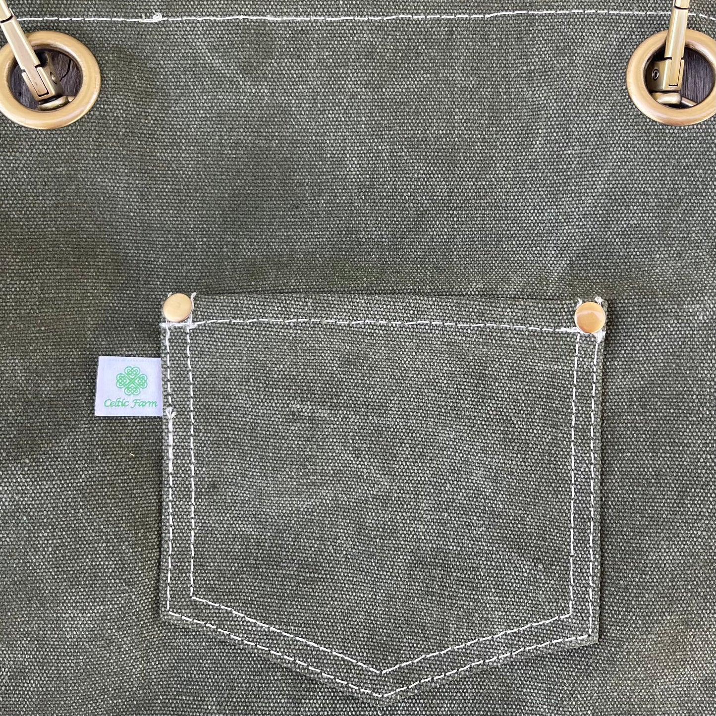 A close-up of The Celtic Farm Gardening Apron shows a green waxed canvas pocket with white stitching, two gold rivets, gold grommets above, and a green logo label—perfect details for this durable Waxed Canvas Apron with Pockets.