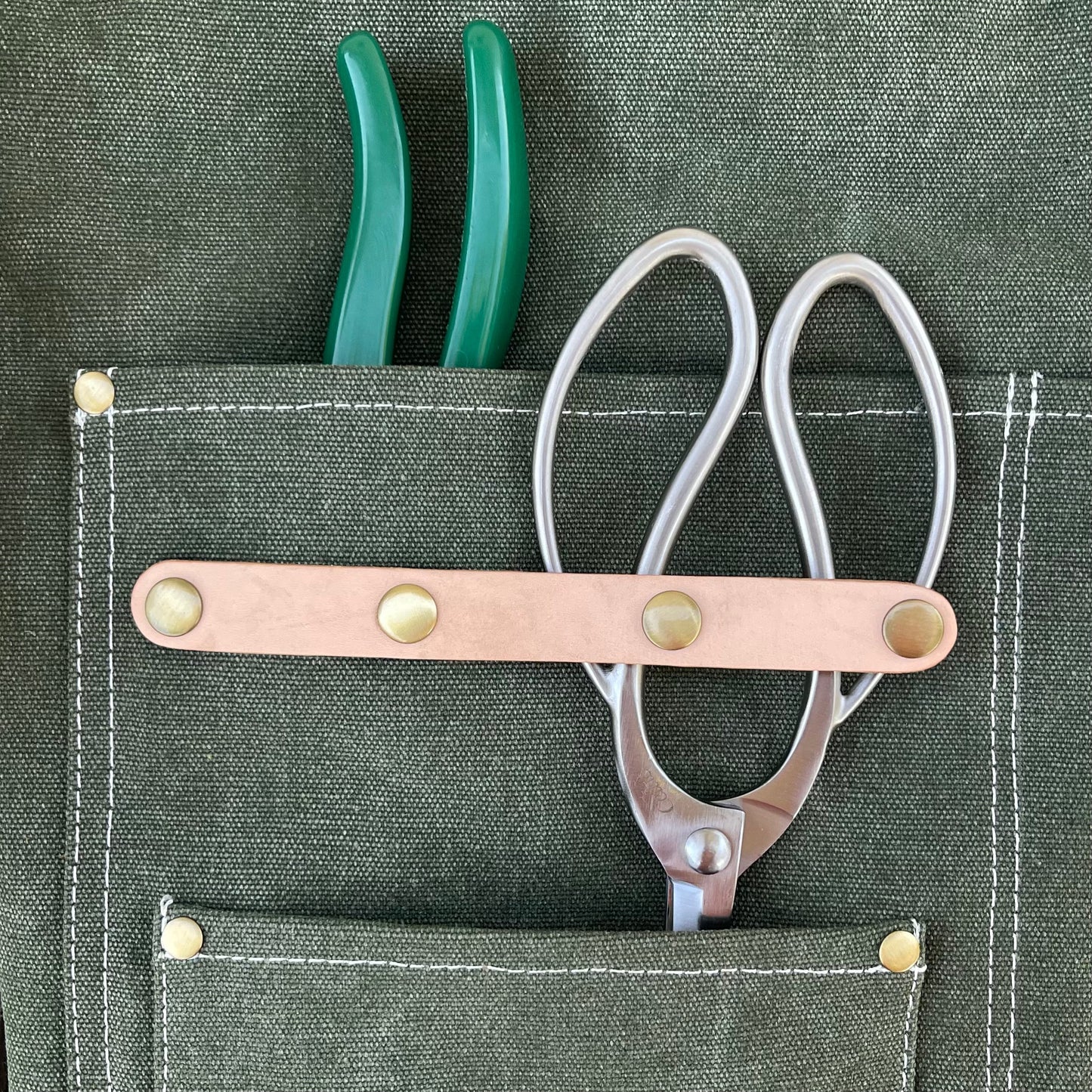 A pair of green-handled pliers and silver scissors with a tan leather strap are tucked into the pockets of The Celtic Farm Gardening Apron—waxed cotton canvas with white stitching and brass rivets.