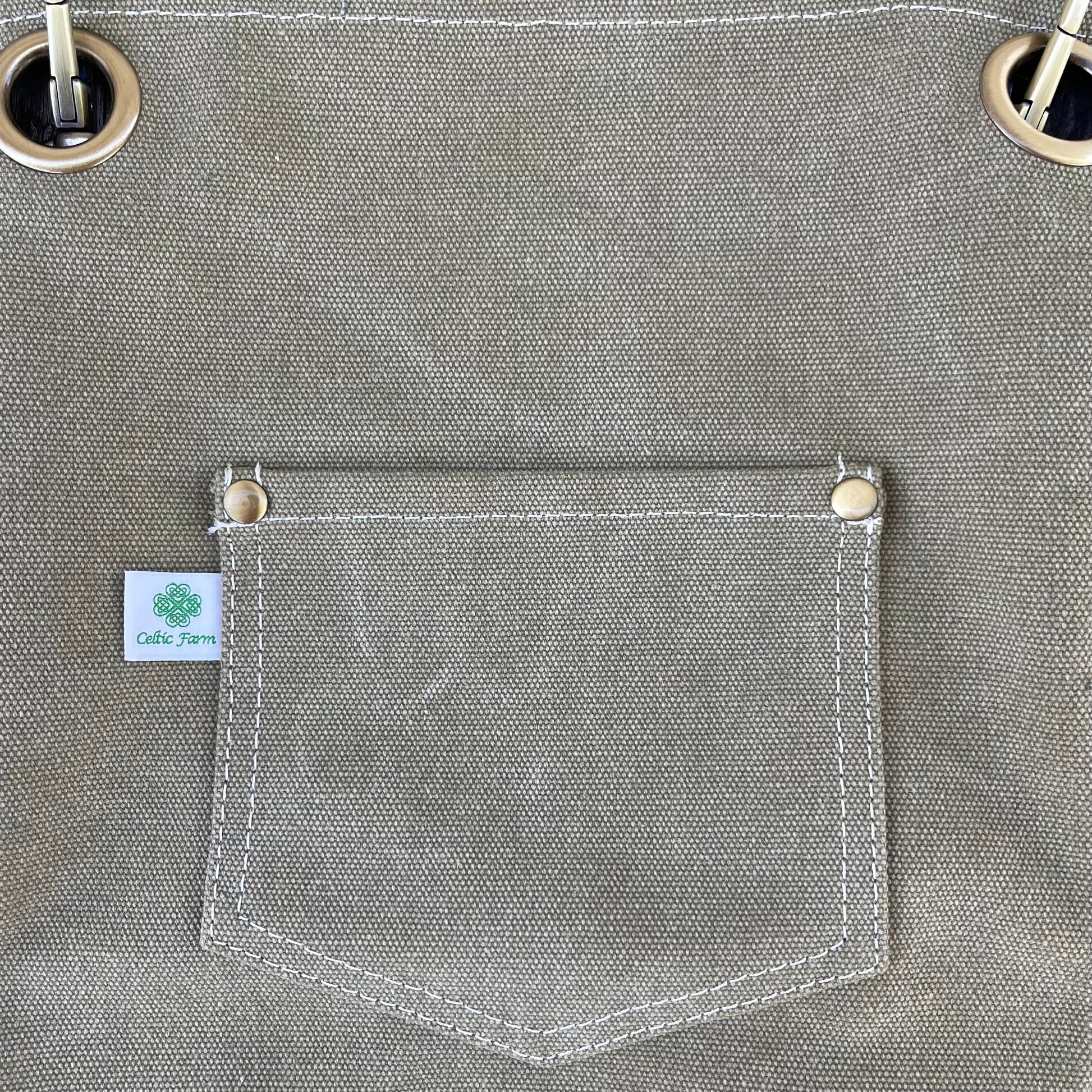 Close-up of The Celtic Farm Gardening Apron—khaki waxed canvas with stitched pocket, white label featuring a green clover, two metal grommets with a white drawstring—durable and perfect for gardening.