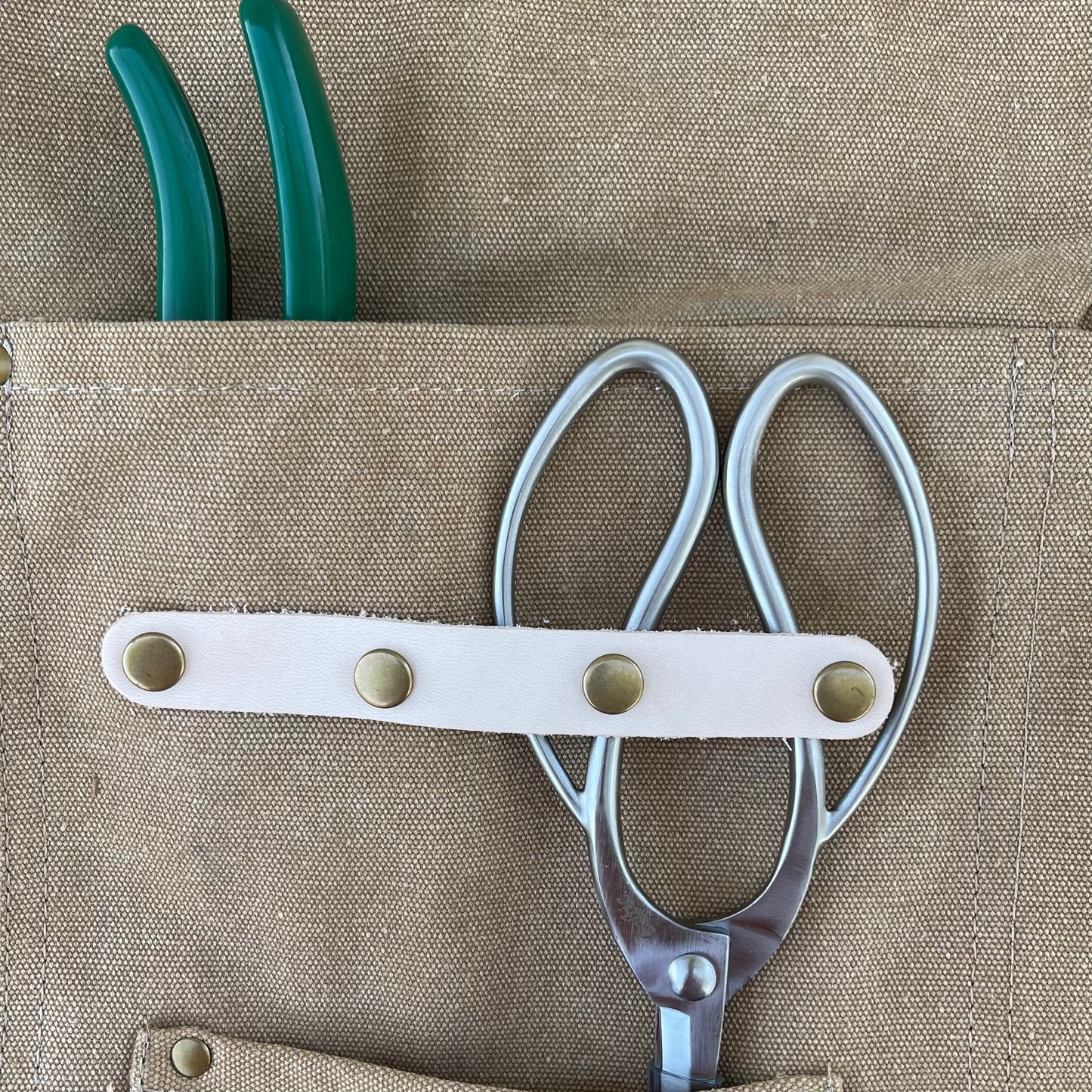 A pair of metal scissors and green-handled pliers sit in the pocket of The Celtic Farm Gardening Apron—Waxed Canvas Apron with Pockets.