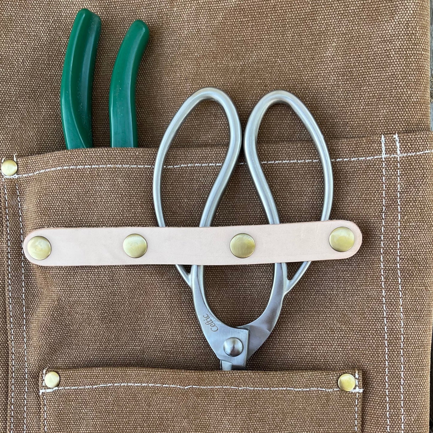 A pair of green-handled pliers and silver scissors are stored in The Celtic Farms Gardening Apron—Waxed Canvas Apron with Pockets, featuring white stitching, brass rivets, and a beige leather strap. Ideal for gardening or florist apron use.