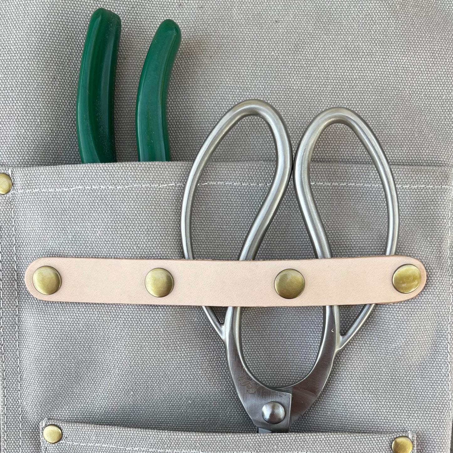 The Celtic Farm’s Gardening Apron—crafted from gray waxed canvas with pockets—holds green-handled pliers and metal scissors, secured by a beige leather strap with brass rivets; perfect for gardeners and florists.